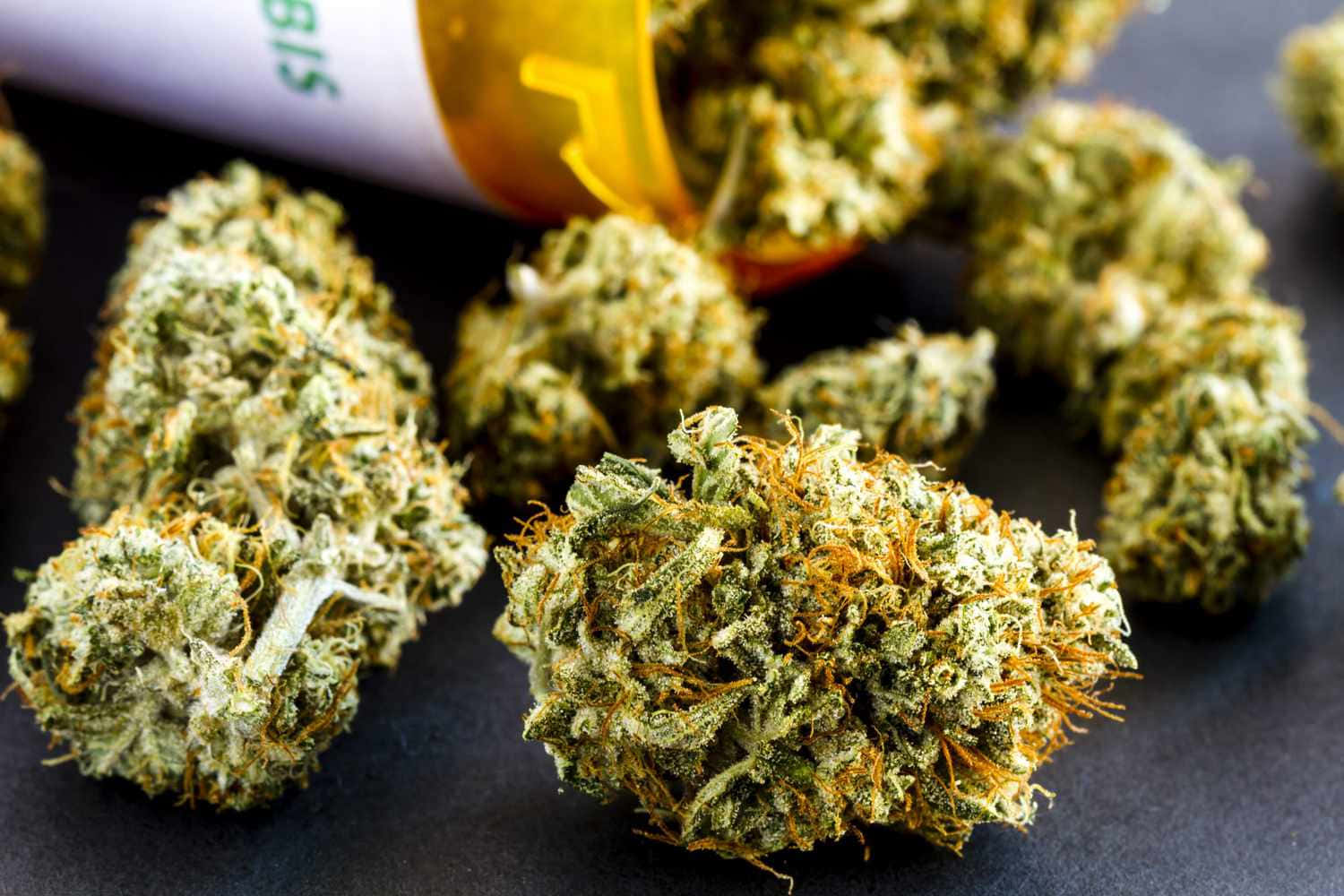 大麻 桌布 壁紙 — 特寫鏡頭：深色表面上，傾斜的琥珀色藥瓶溢出濃密的綠色大麻花蕾，帶有橙色雌蕊。