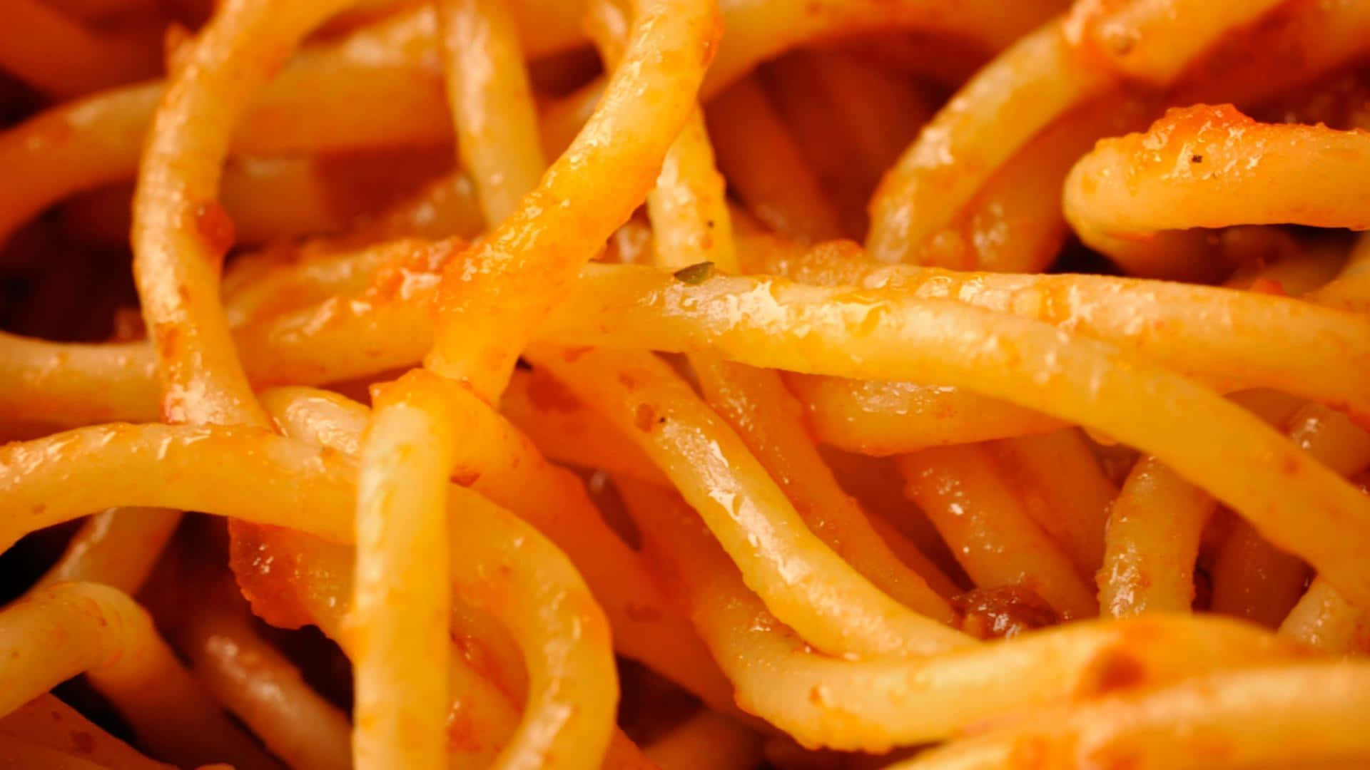 Pasta 桌布 壁紙 — 特寫鏡頭：濃郁番茄醬包裹的亮澤義大利麵，可見香草和油在溫暖燈光下閃爍。