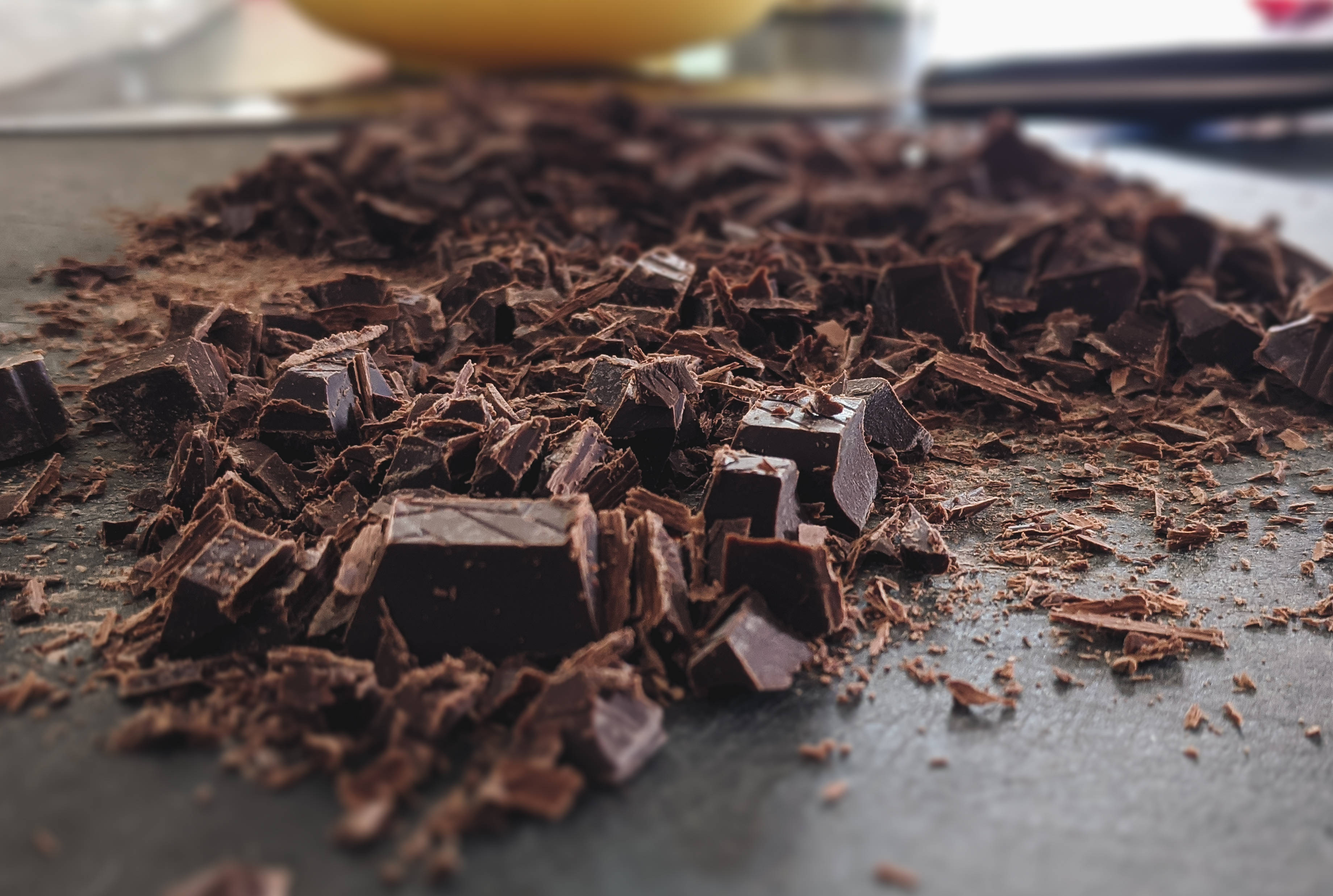 Chocolate 桌布 壁紙 — 特寫鏡頭：濃郁黑巧克力塊和碎屑散落在深色表面上，背景有一個模糊的黃色物體。