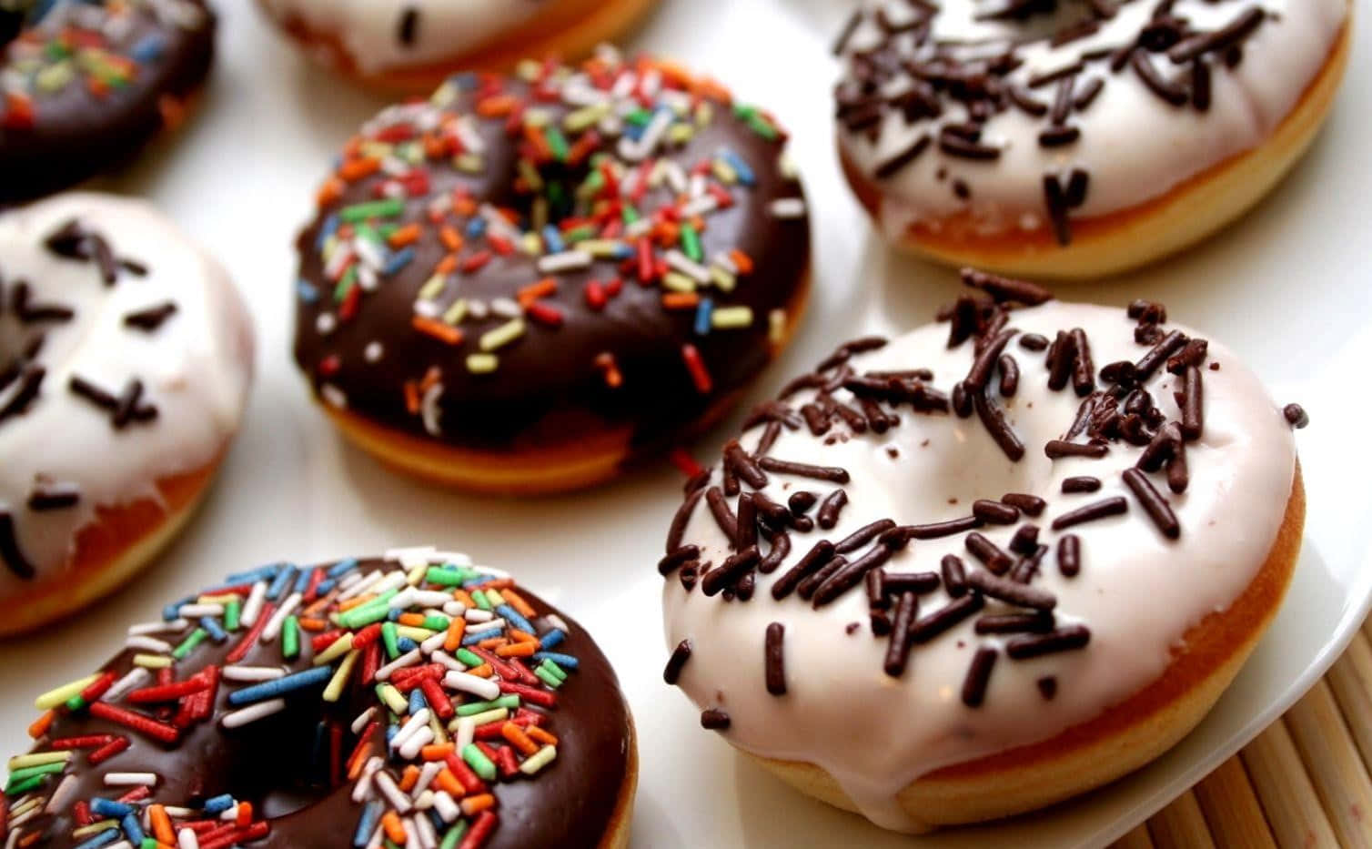 Donut 桌布 壁紙 — 特寫鏡頭：白色盤子上放有各式各樣的糖霜甜甜圈，上面綴有彩色糖粒和巧克力醬。