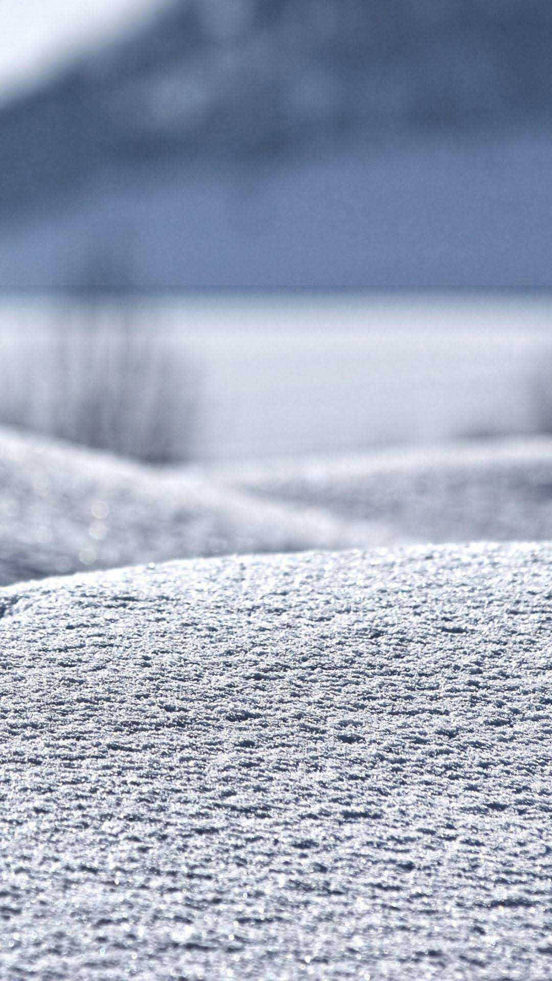 iPhone 桌布 壁紙 — 特寫鏡頭：質感豐富的雪堆在柔和光線下閃閃發光，背景是模糊的雪山。