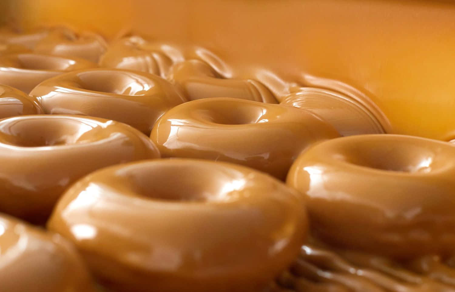 Glazed Donut 桌布 壁紙 — 特寫鏡頭：閃亮焦糖釉面甜甜圈，排列成溫暖金色調，搭配柔和散景背景。