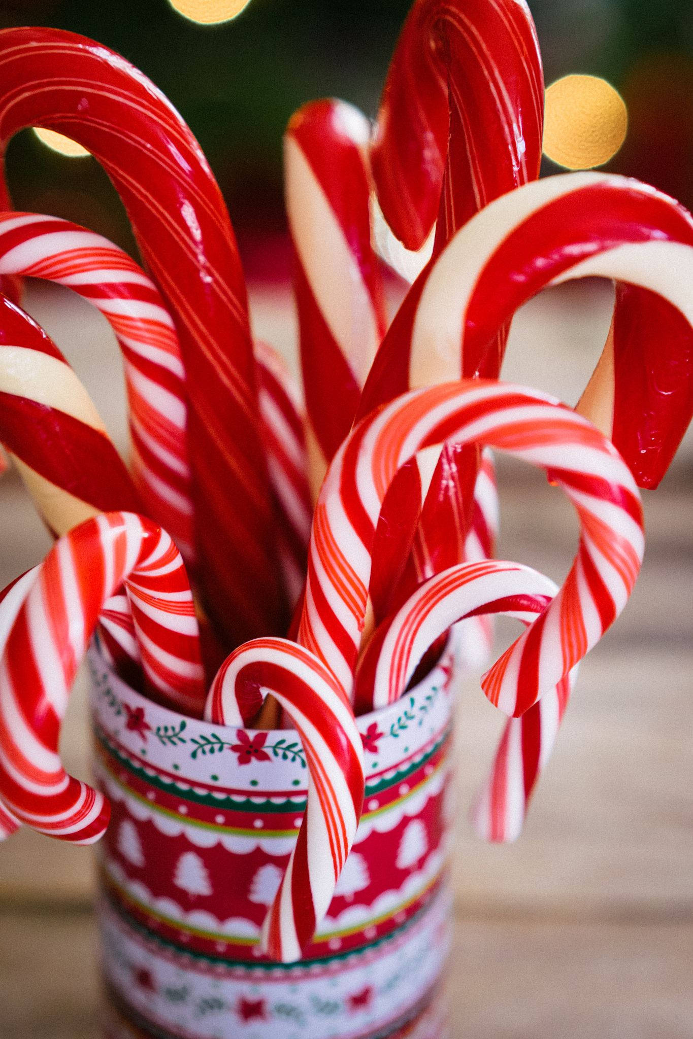 Candy Cane 桌布 壁紙 — 特寫鏡頭，閃亮的紅白條紋拐杖糖從節日圖案杯子中溢出，背後有模糊的溫暖燈光。