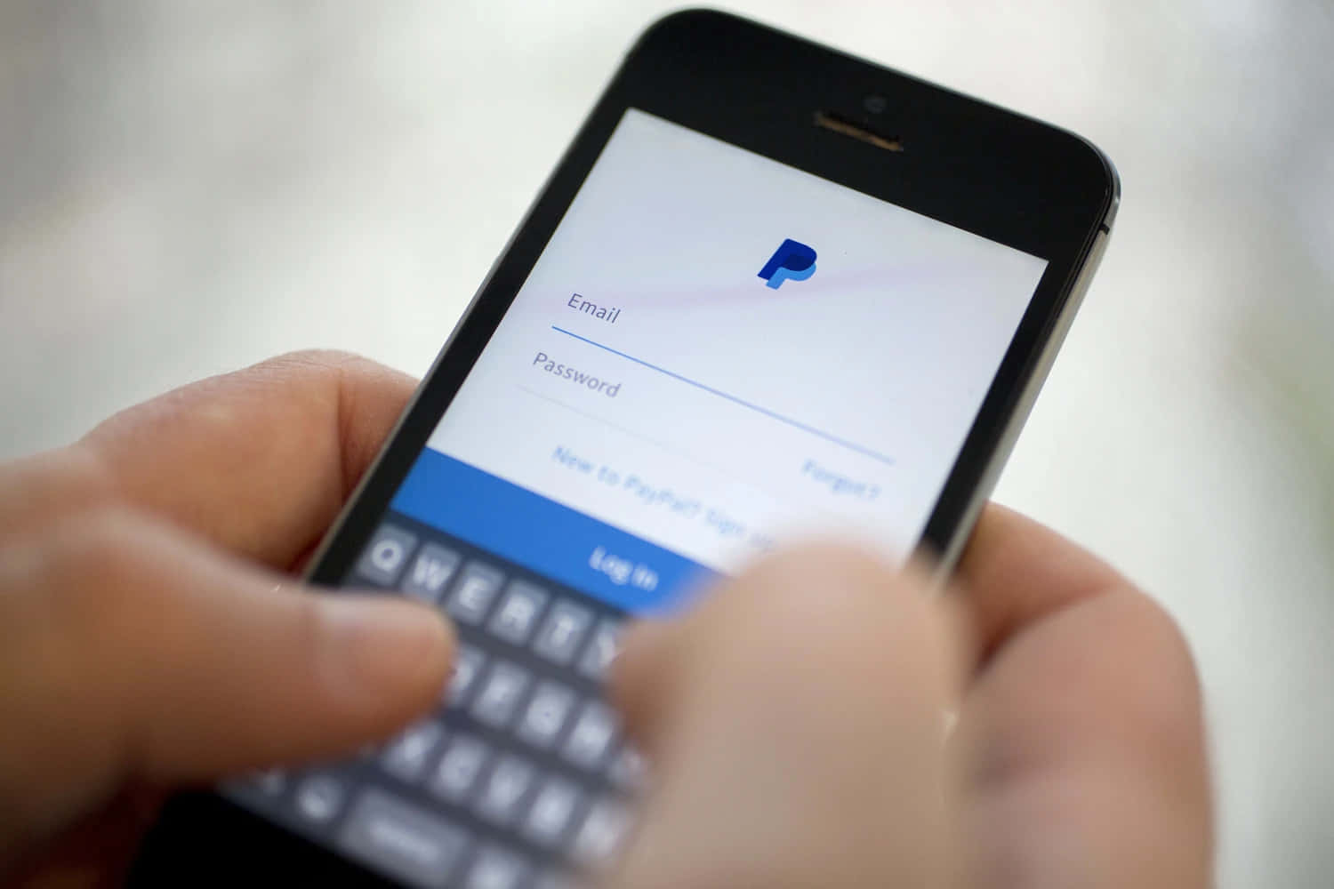 PayPal 桌布 壁紙 — 特寫鏡頭：雙手在模糊背景的智慧型手機上輸入PayPal應用程式的登入憑證。