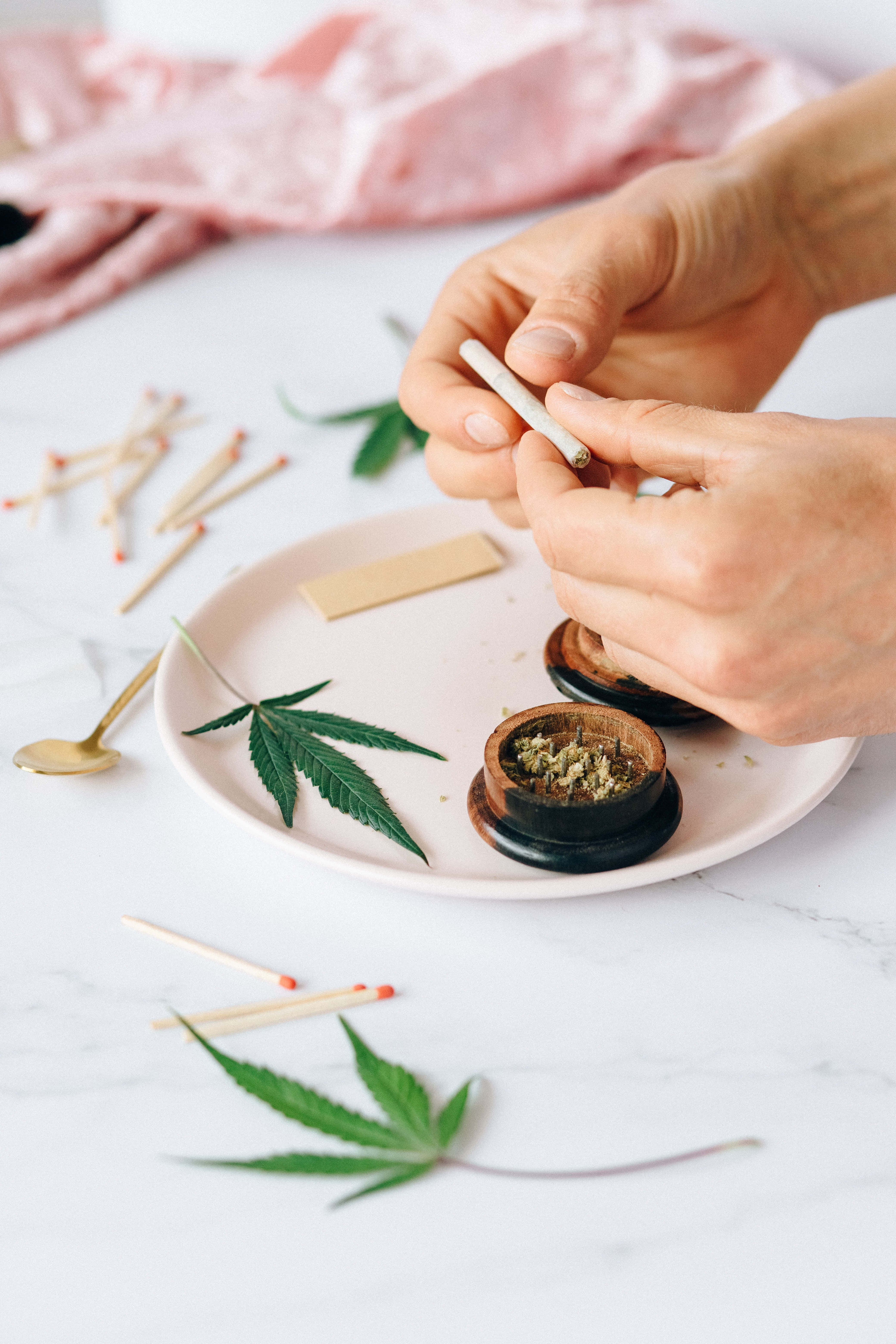 Weed Blunt 桌布 壁紙 — 特寫鏡頭，雙手在白色大理石表面上捲大麻煙，周圍散落著綠葉和火柴。