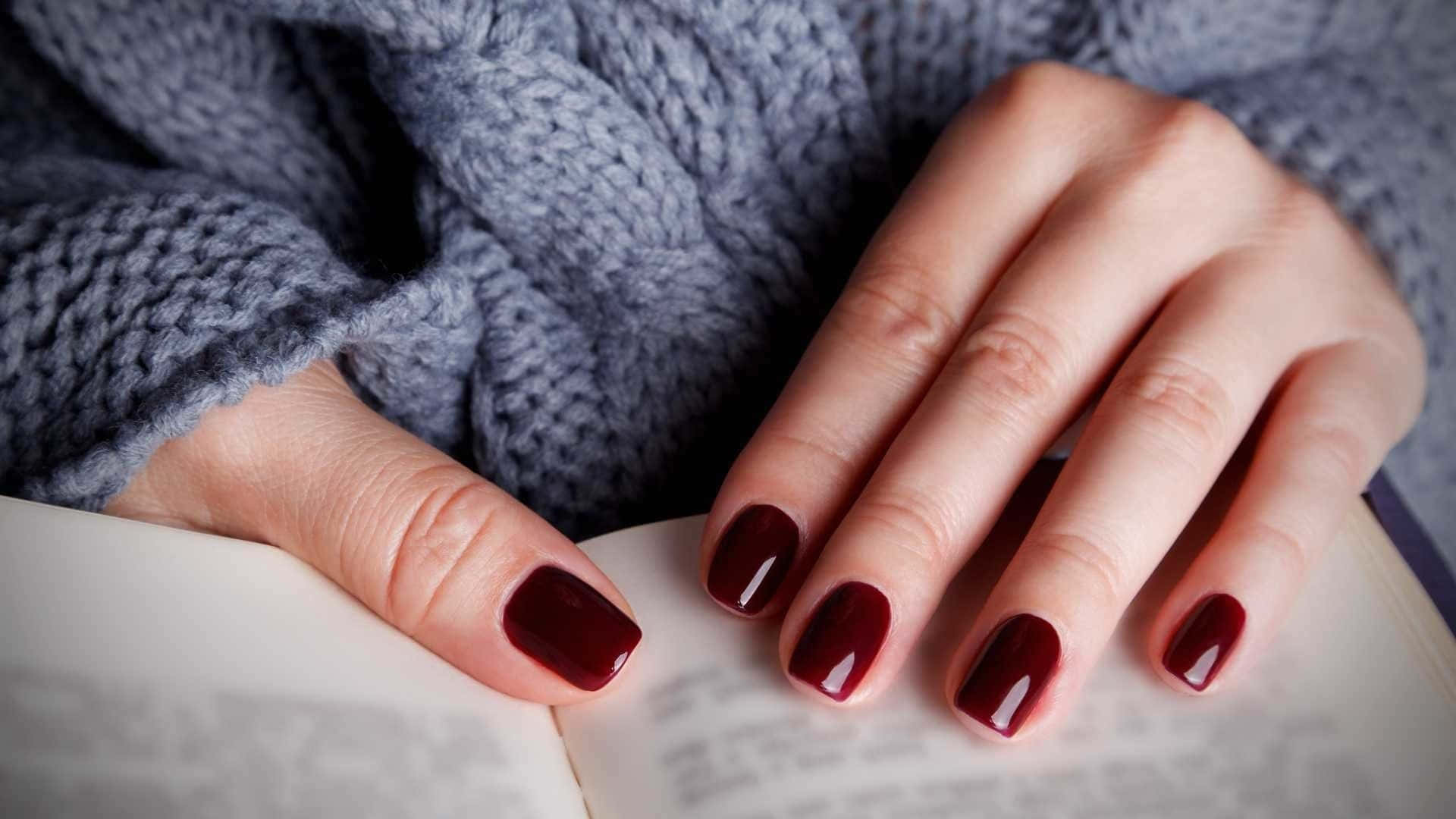 Nail 桌布 壁紙 — 特寫鏡頭：雙手塗有光澤酒紅色指甲油，擱在一本打開的書上，背景是質感灰色毛衣。
