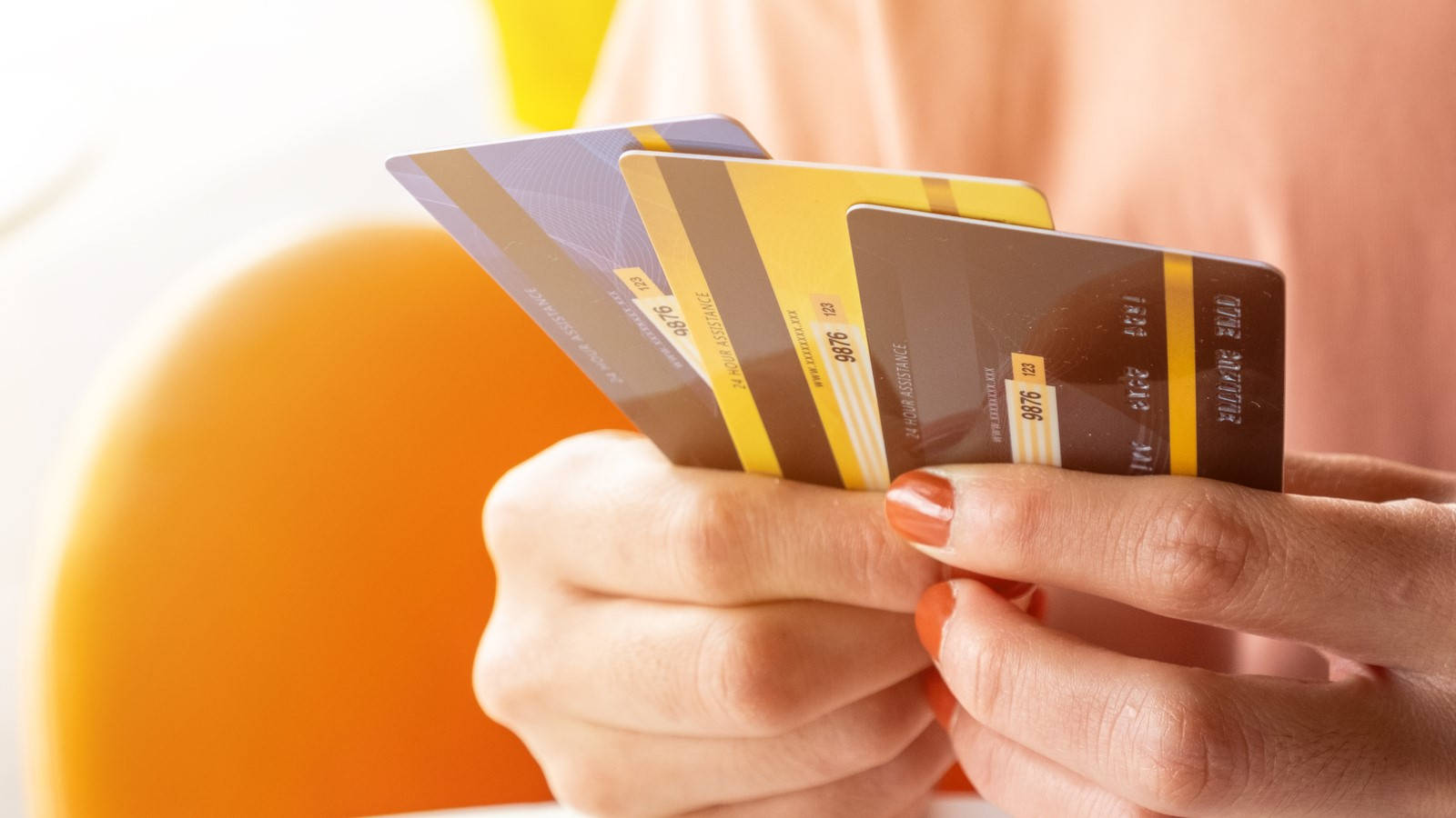 Credit Card 桌布 壁紙 — 特寫鏡頭，雙手拿著多張信用卡，塗有橙色指甲油，背景是模糊的橙色椅子。