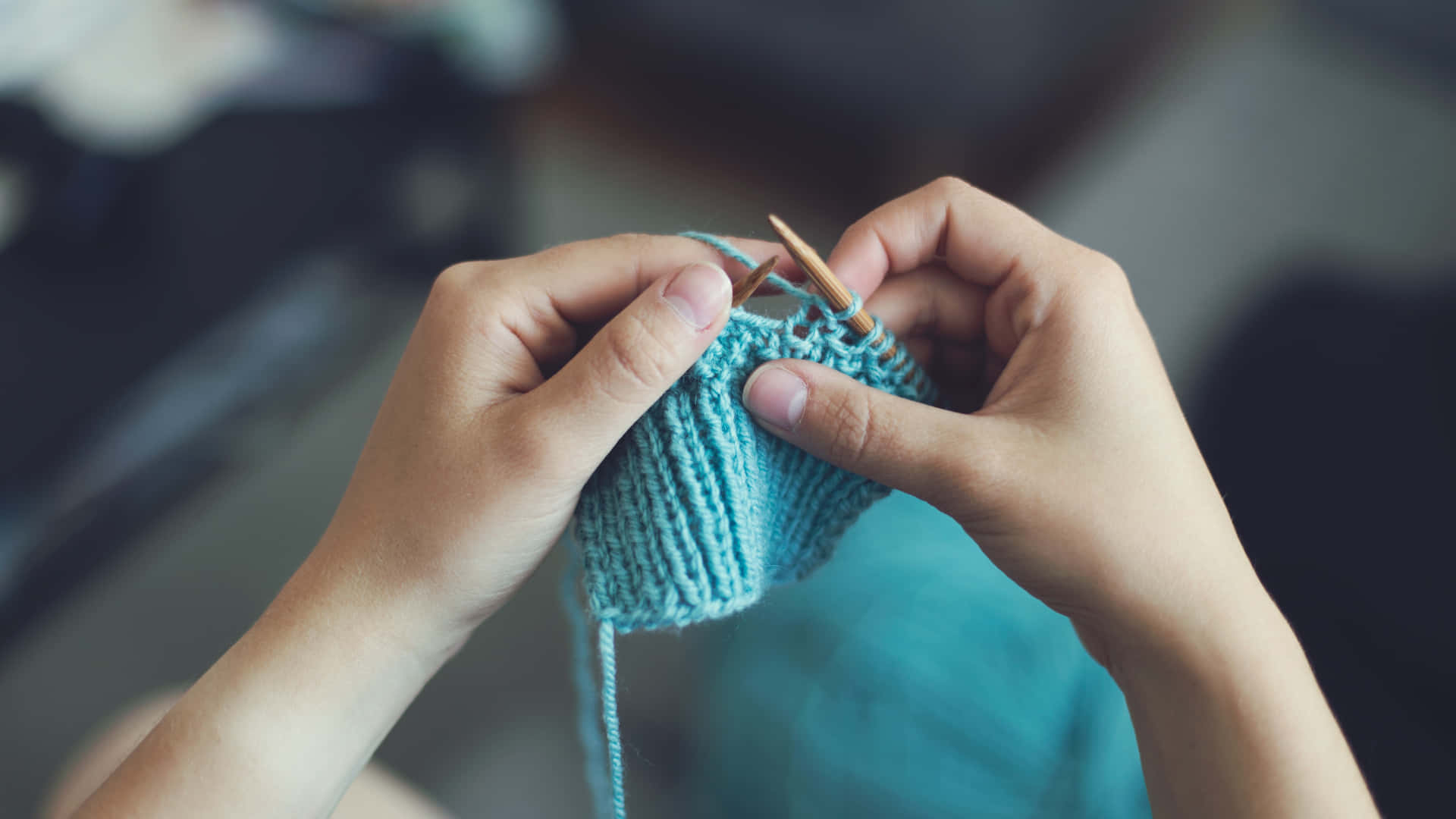 Knitting 桌布 壁紙 — 特寫鏡頭：雙手用木針編織藍綠色布料，背景為模糊的室內環境。