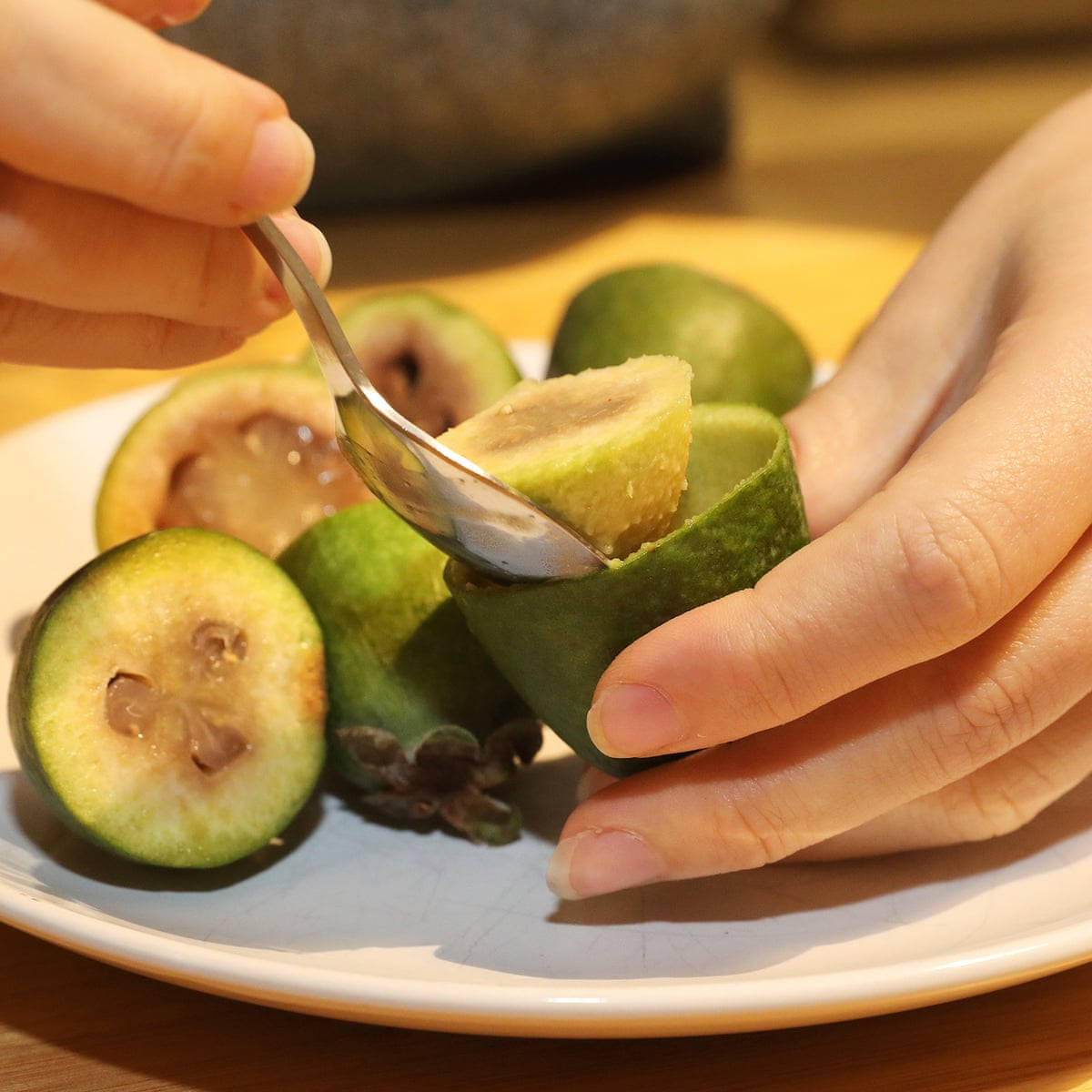 Fruit 桌布 壁紙 — 特寫鏡頭：雙手用湯匙從白色盤子上的半個綠色菲油果中舀出甜美的果肉。