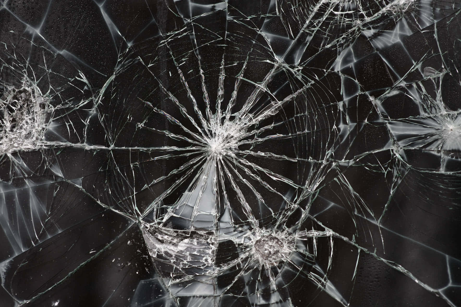 Broken Glass 桌布 壁紙 — 特寫鏡頭，顯示一塊破碎玻璃，在深色背景下，撞擊點周圍有複雜的裂紋向外擴散。