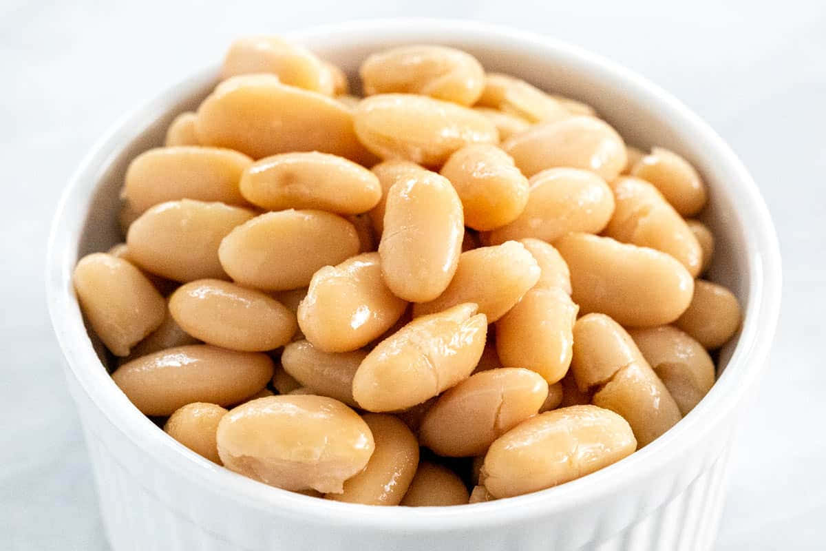 beans 桌布 壁紙 — 特寫鏡頭：飽滿的奶油白豆在白色陶瓷碗中閃閃發光，襯以柔和淺色背景。