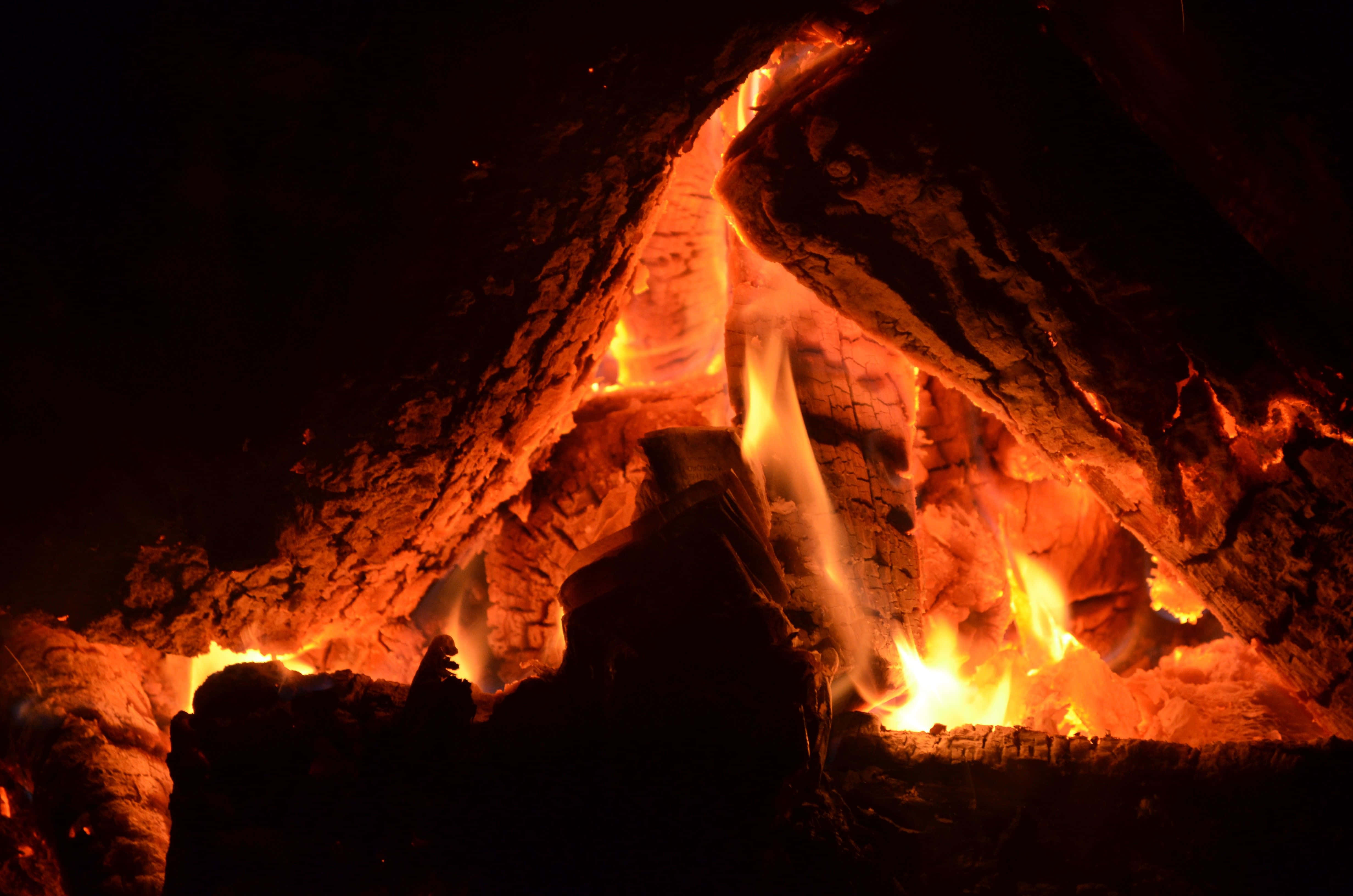 Campfire 桌布 壁紙 — 特寫鏡頭：黑暗壁爐中發光的餘燼和劈啪作響的火焰，將溫暖的橙色光芒投射在燒焦的木頭上。