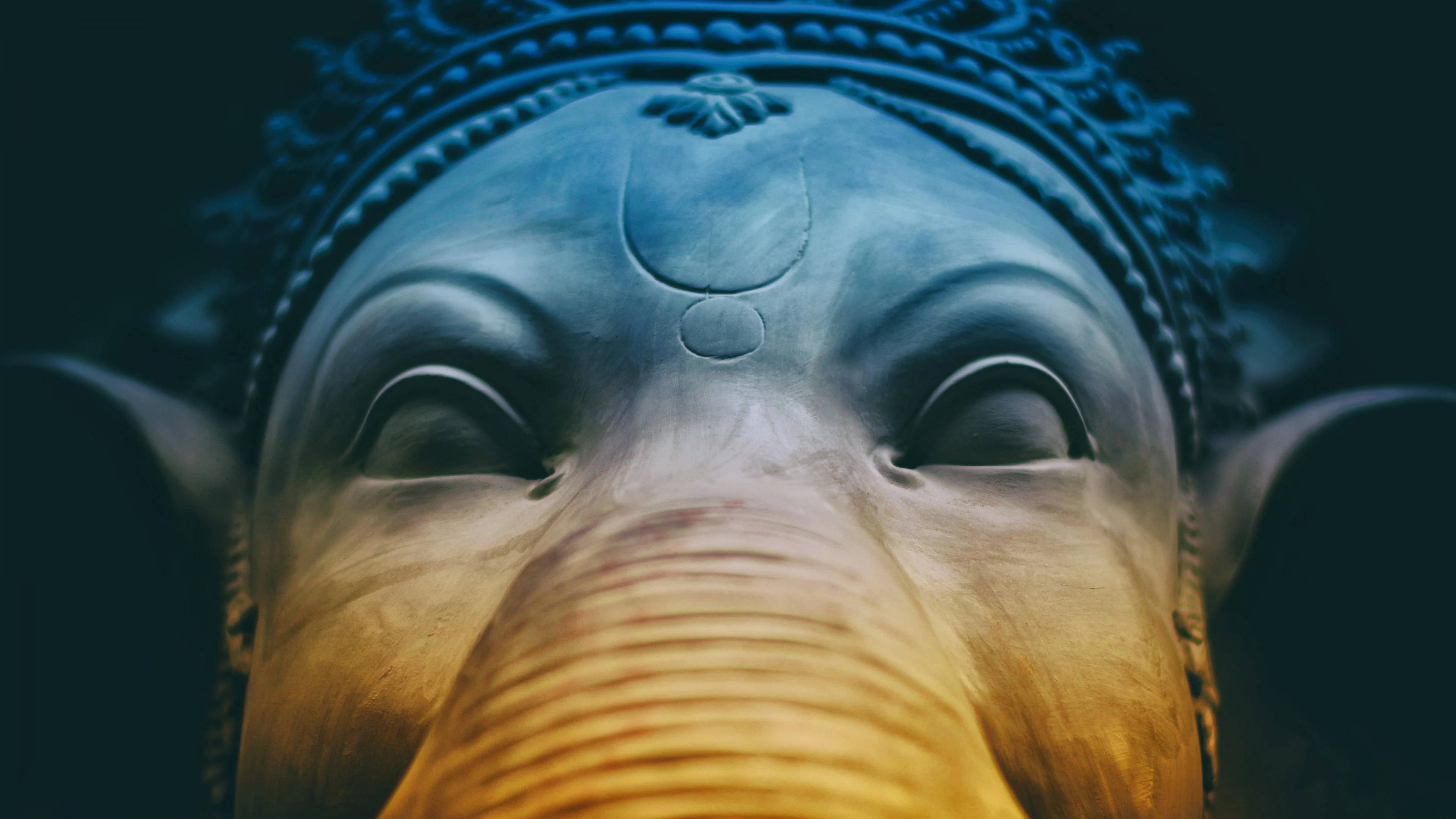 ganesh 4K 桌布 壁紙 — 特寫鏡頭下，一尊寧靜的象頭雕像，戴著藍色皇冠和金色象鼻，在戲劇性燈光中呈現。