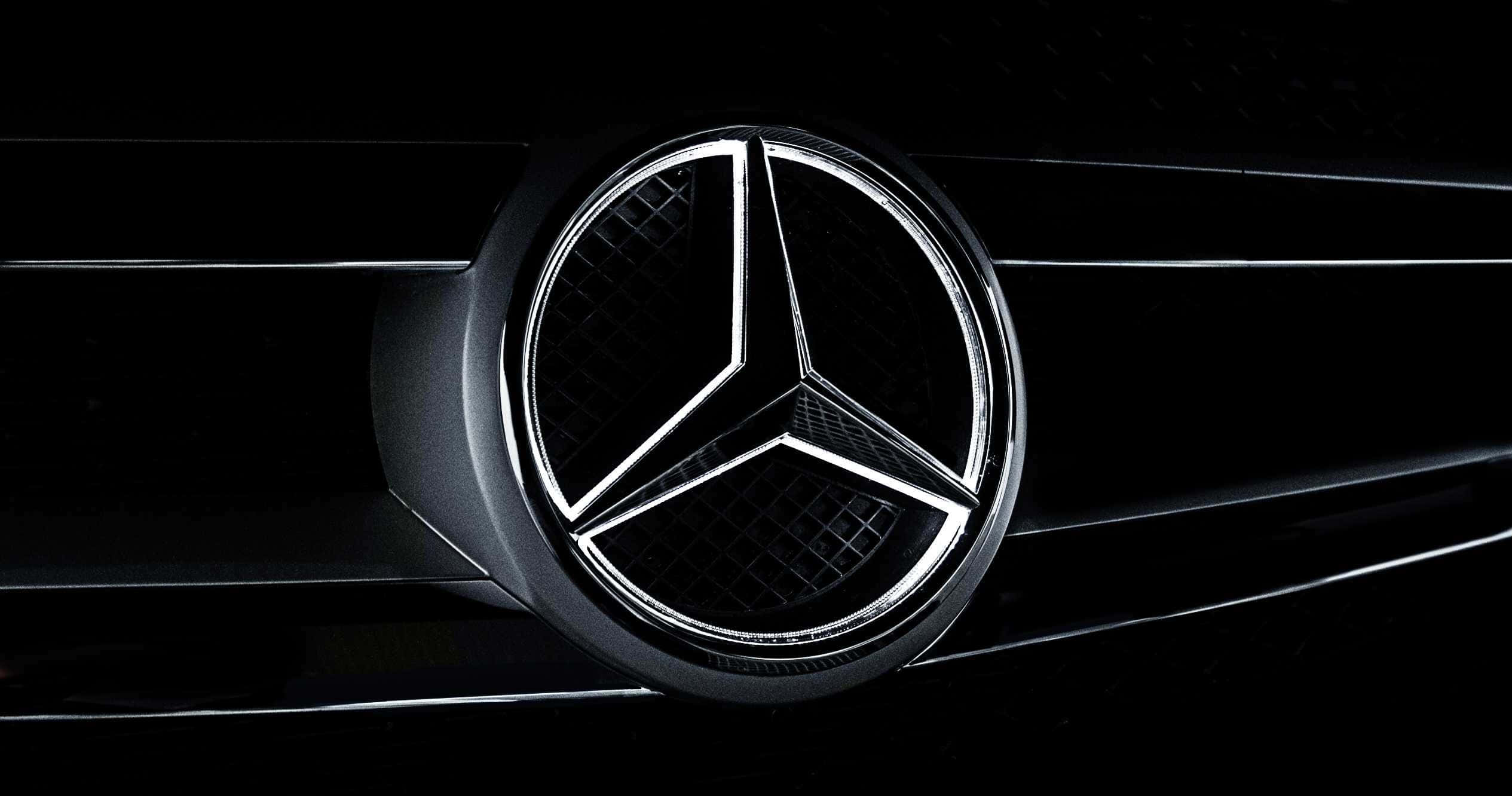 Car Logo 桌布 壁紙 — 特寫鏡頭下，黑色汽車格柵中央閃耀的梅賽德斯-奔馳三叉星徽標，在戲劇性燈光中呈現。