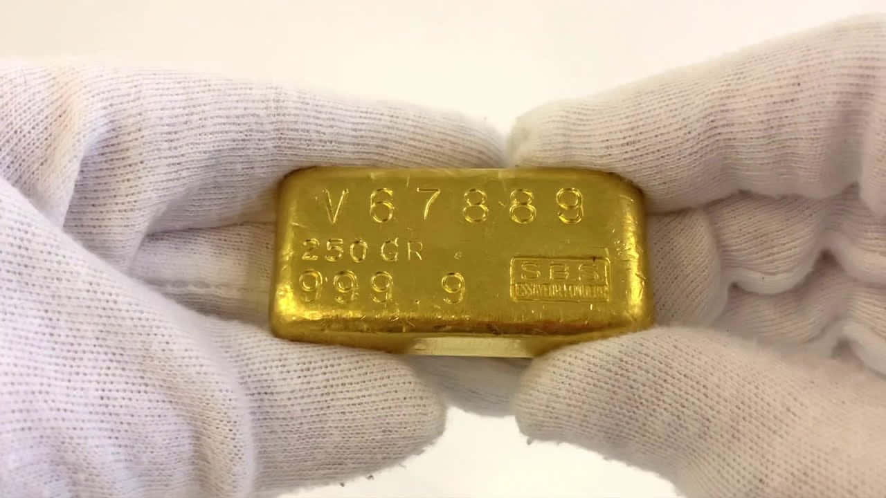 Gold Bar 桌布 壁紙 — 特寫鏡頭展示一隻戴手套的手拿著一塊250克金條，突顯其雕刻標記和閃亮的金屬表面。