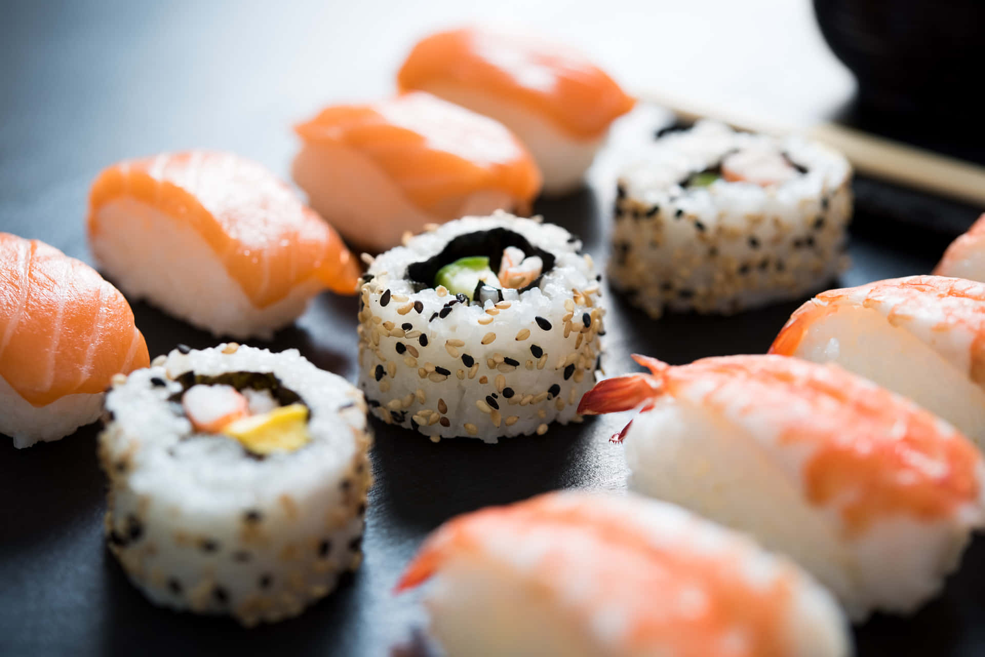 Sushi 桌布 壁紙 — 特寫鏡頭展示各種壽司，包括鮭魚握壽司和芝麻卷，排列在深色盤子上。