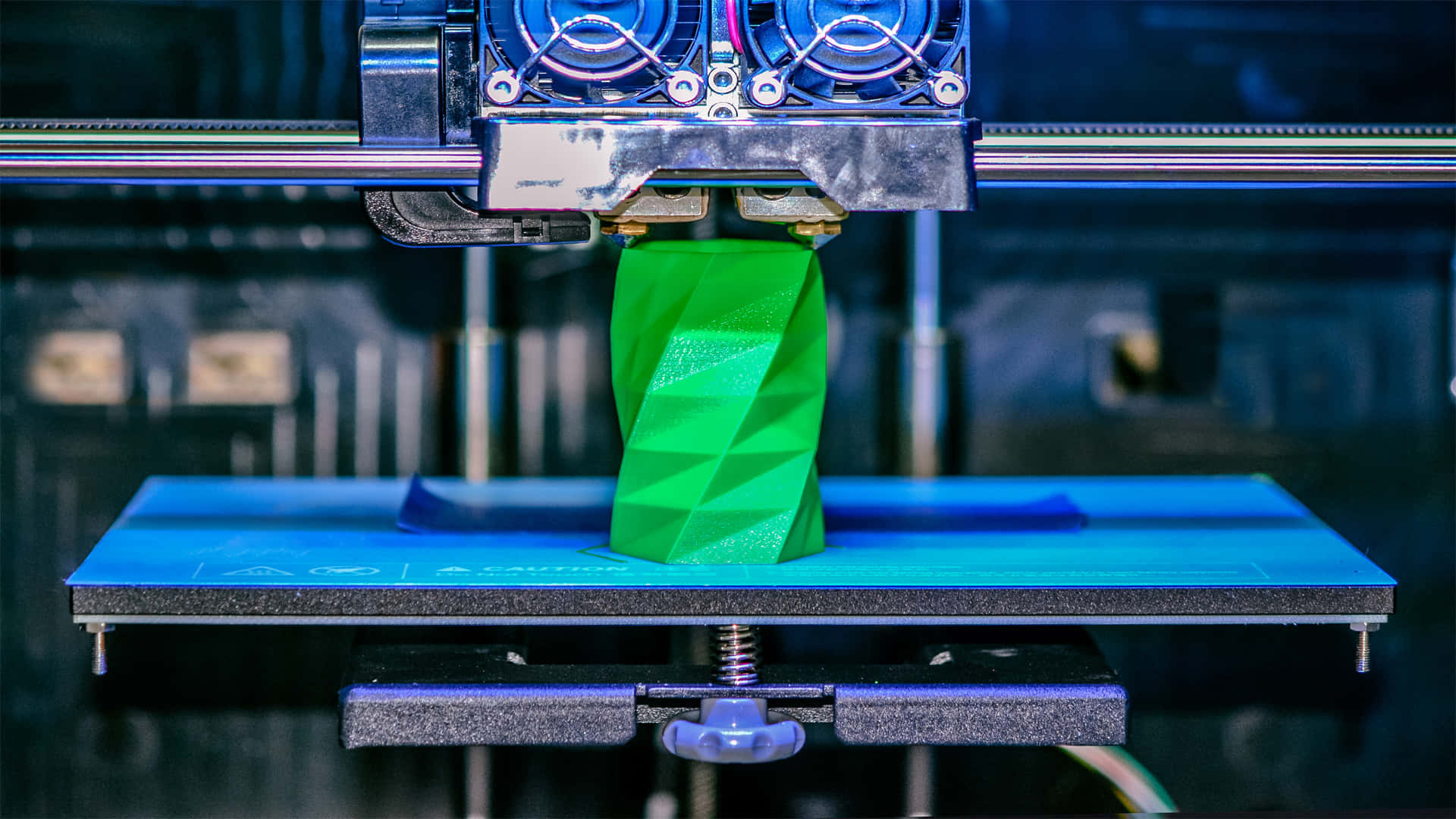 3d Printer 桌布 壁紙 — 特寫鏡頭顯示3D列印機在藍色列印平台上擠出鮮綠色線材，構建幾何螺旋物體。