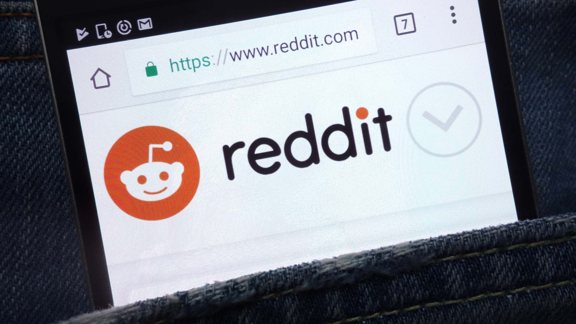 reddit 桌布 壁紙 — 特寫鏡頭顯示一支智慧型手機，螢幕上展示Reddit首頁，標誌和網址清晰可見，手機放在牛仔褲口袋中。