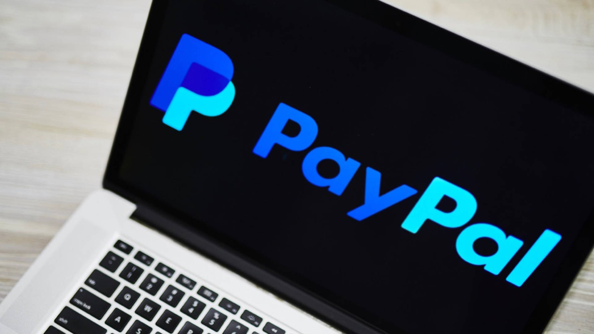 PayPal 桌布 壁紙 — 特寫鏡頭顯示一部筆記型電腦，在深色螢幕上展示PayPal標誌，背景為淺色木桌。