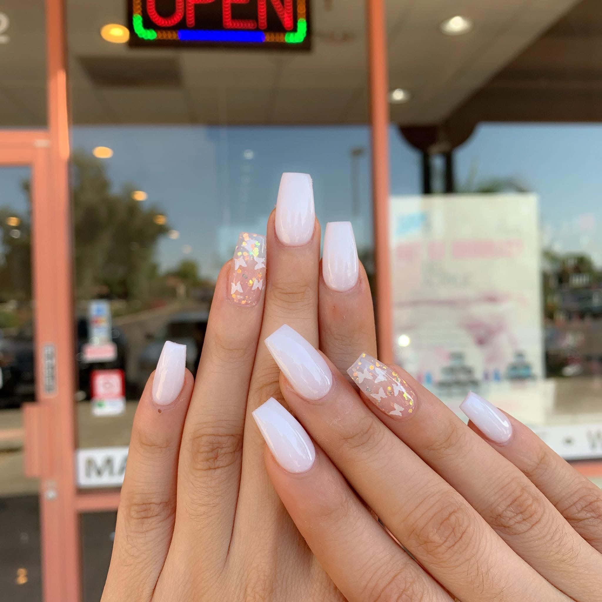 Nail Polish 桌布 壁紙 — 特寫雙手，帶有光澤白色棺材形指甲，飾有亮粉和蝴蝶圖案，背景為模糊的店舖門面。