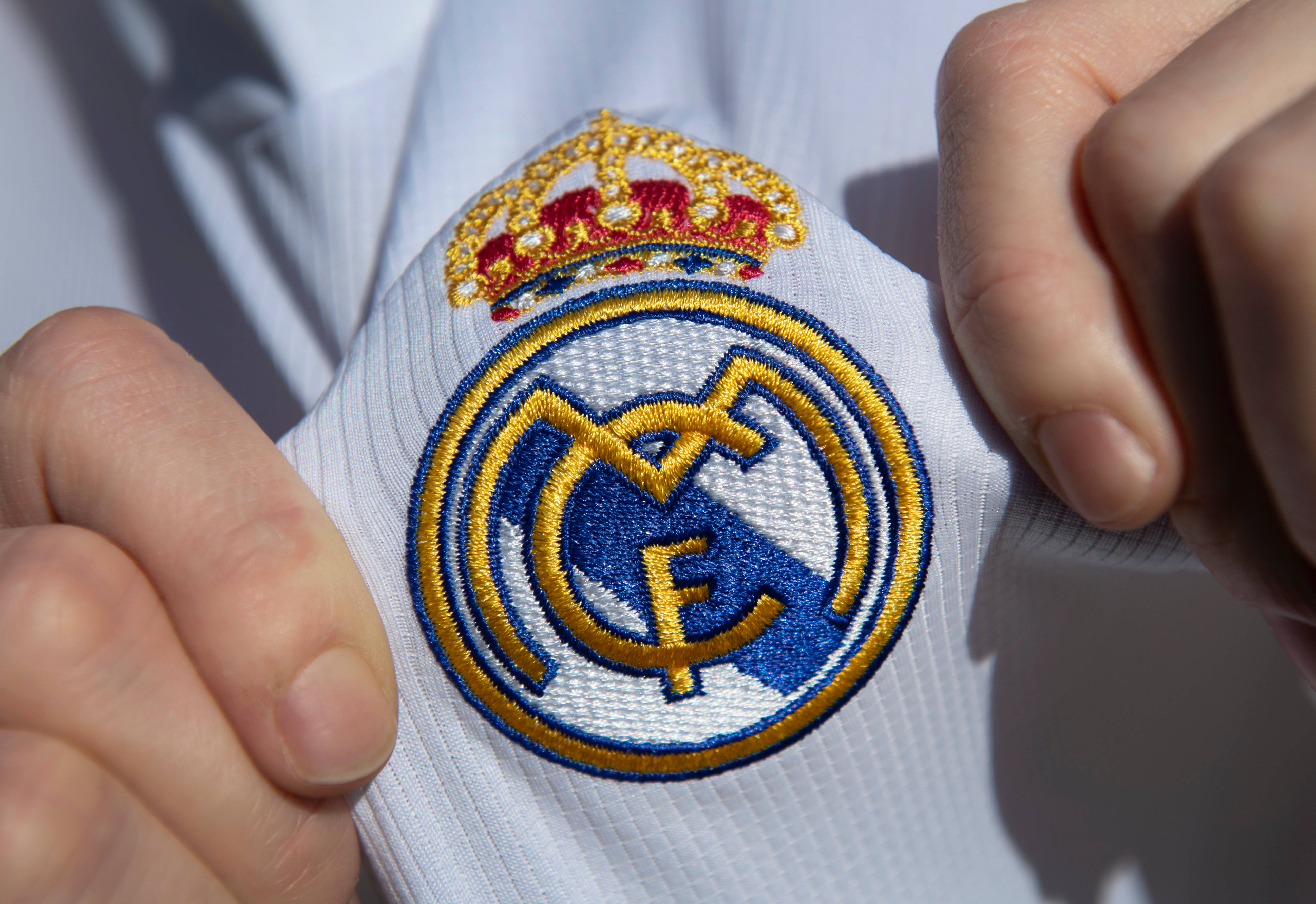 Real Madrid 4K 桌布 壁紙 — 特寫雙手捧著一件白色球衣，展示以金、藍、紅線精緻刺繡的皇家馬德里會徽。