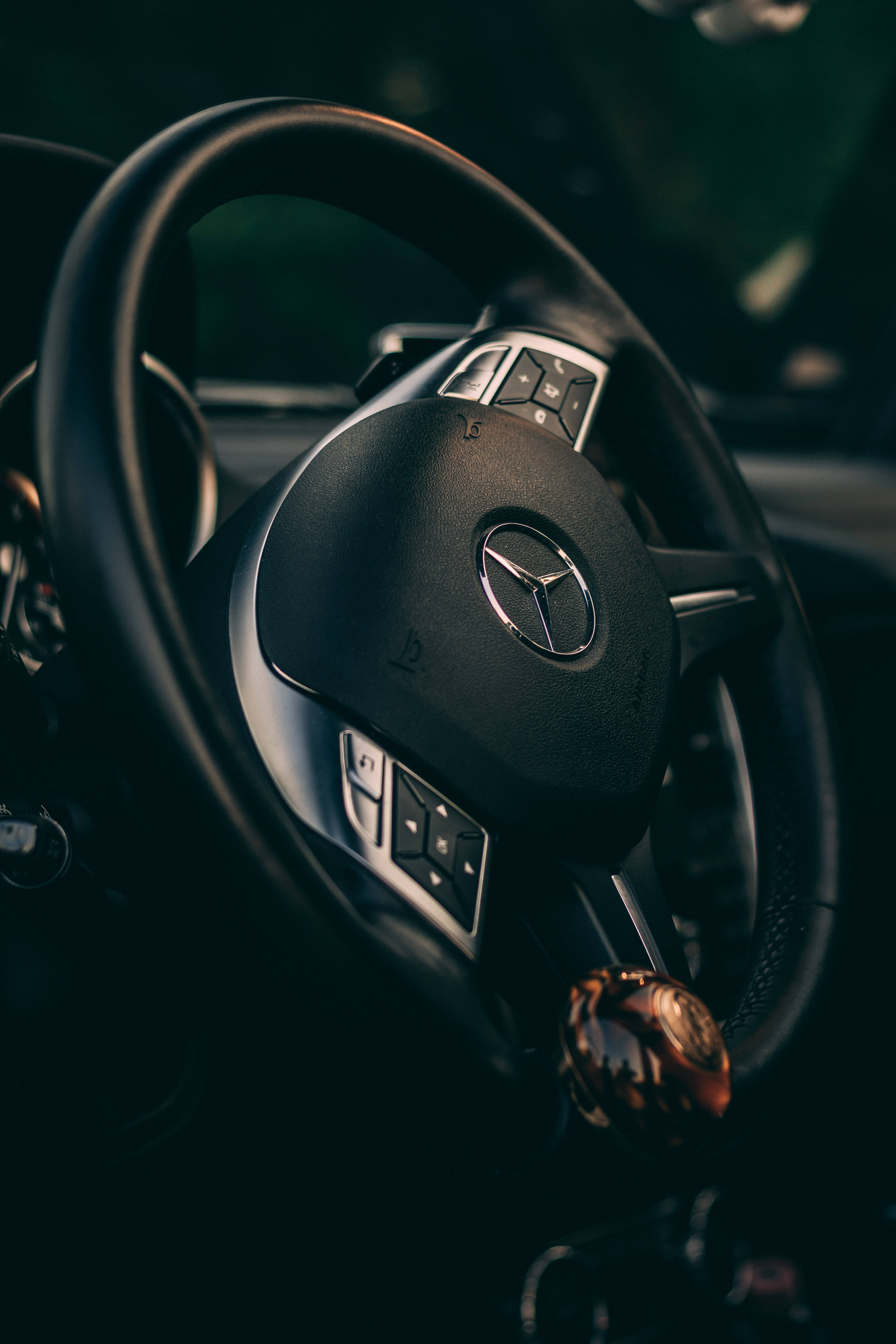 高畫質 賓士 桌布 壁紙 — 特寫黑色 Mercedes-Benz 方向盤，帶有鍍鉻控制裝置和掛在輪輻上的手錶，光線昏暗。
