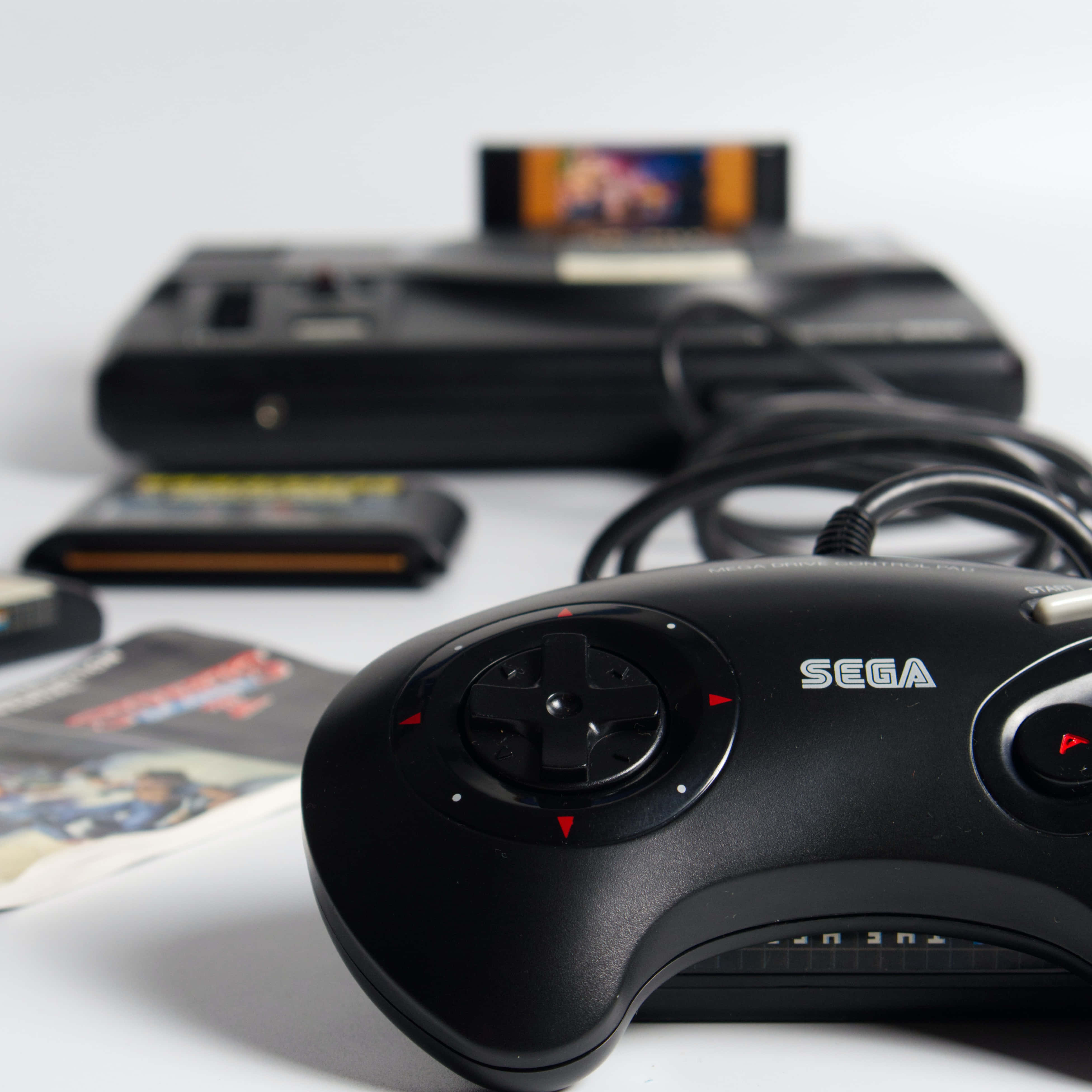 Gaming Consoles 桌布 壁紙 — 特寫黑色Sega Genesis控制器，紅色方向箭頭清晰對焦，背景中的主機和遊戲卡帶模糊。