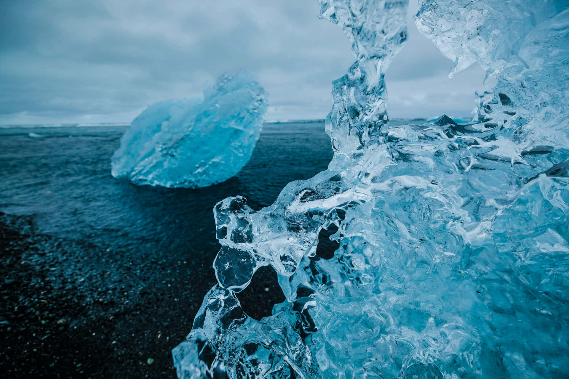 climate change 桌布 壁紙 — 特寫黑色火山海灘上參差不齊的藍色冰層，遠處有浮冰，天空多雲。