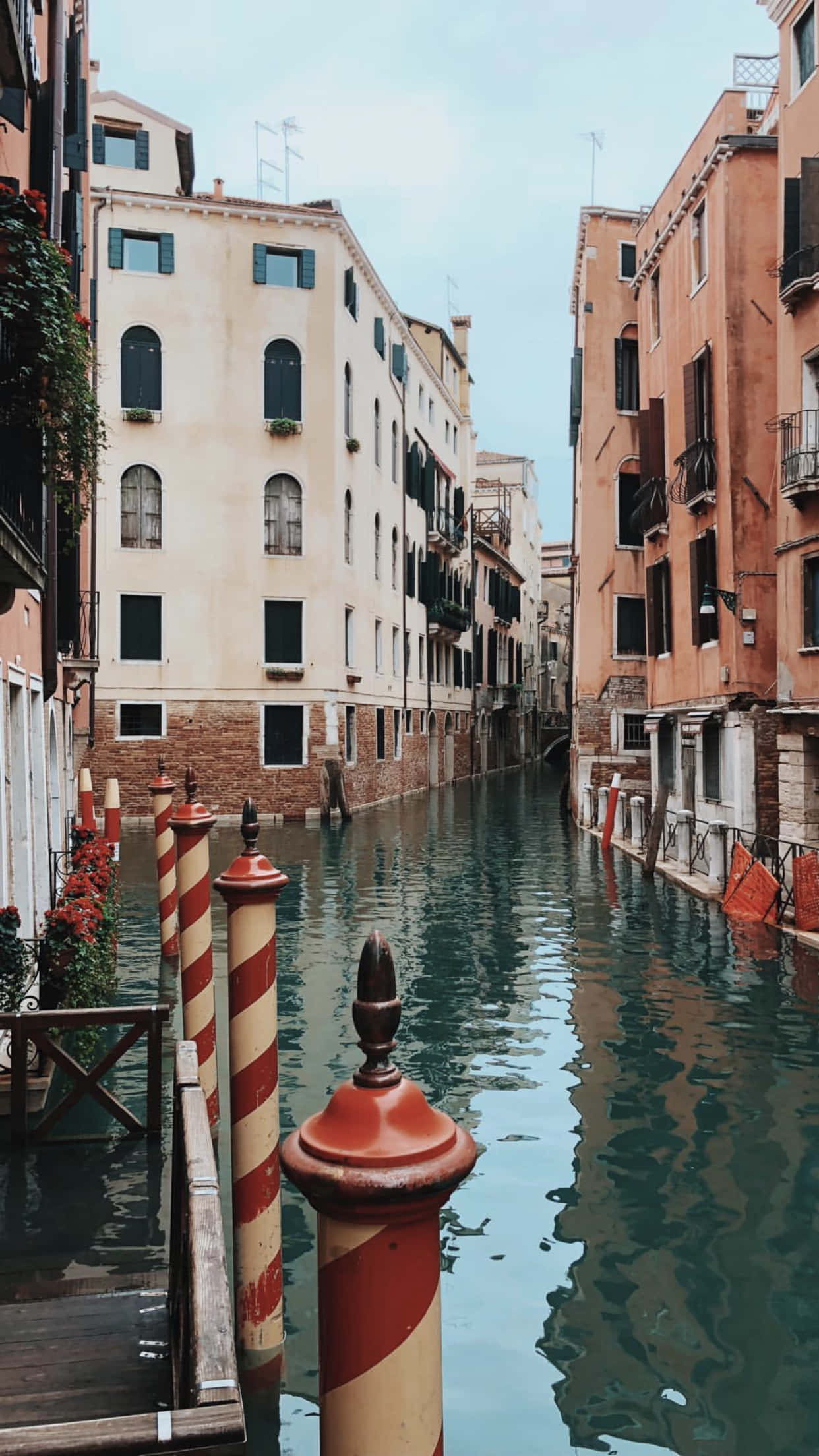Italy Aesthetic 桌布 壁紙 — 狹窄的威尼斯運河兩旁是歷史建築，平靜的水面上有紅白條紋的柱子，天空多雲。