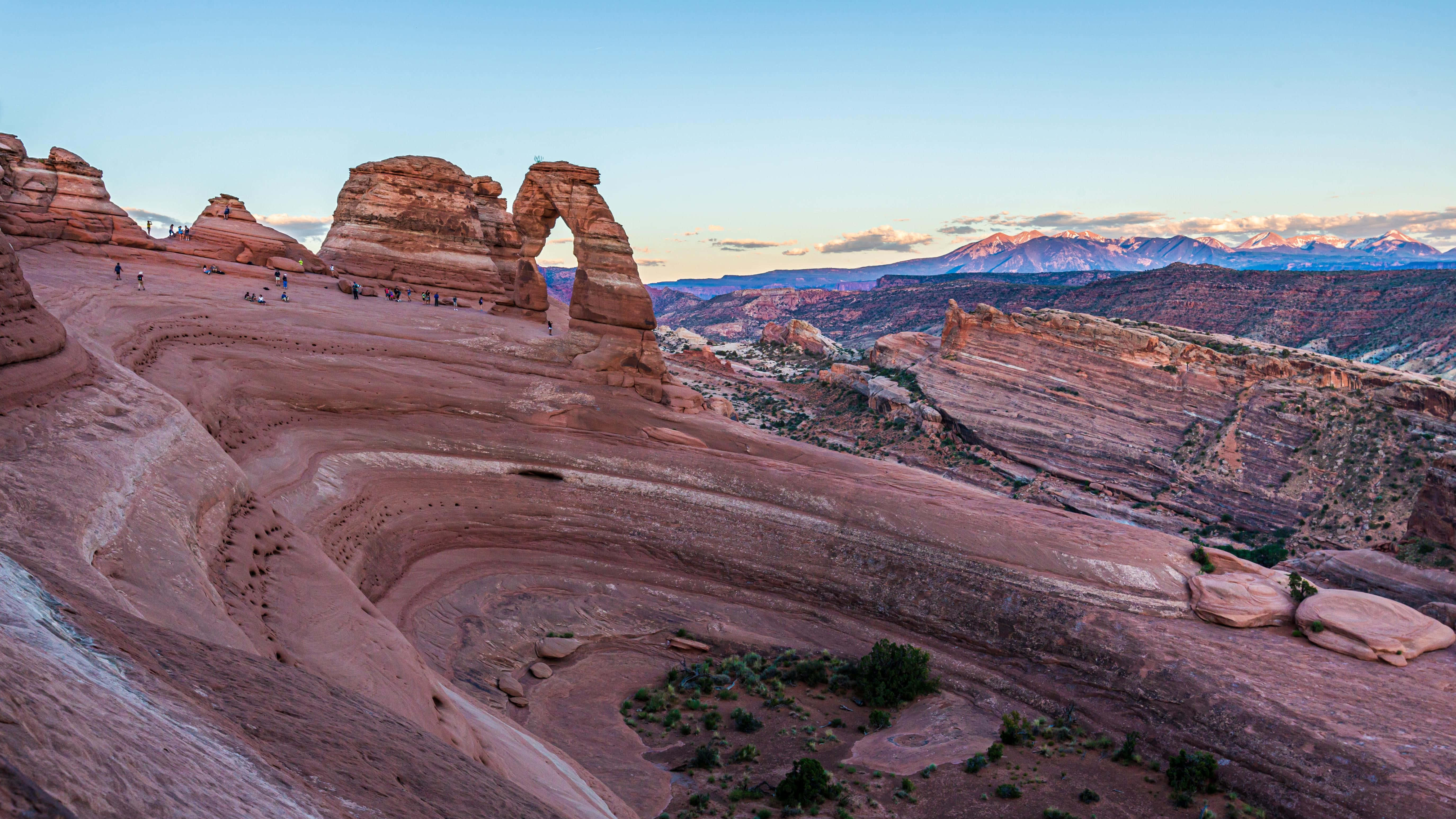 Rock 桌布 壁紙 — 猶他州精緻拱門的壯麗日落景色，步道上有徒步旅行者，遠處有白雪覆蓋的山脈。