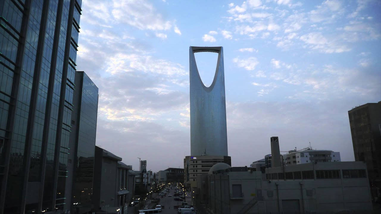 Riyadh 桌布 壁紙 — 王國塔在柔和的暮色天空與散落的雲朵襯托下宏偉聳立，兩旁是現代玻璃摩天大樓和下方的城市街道。