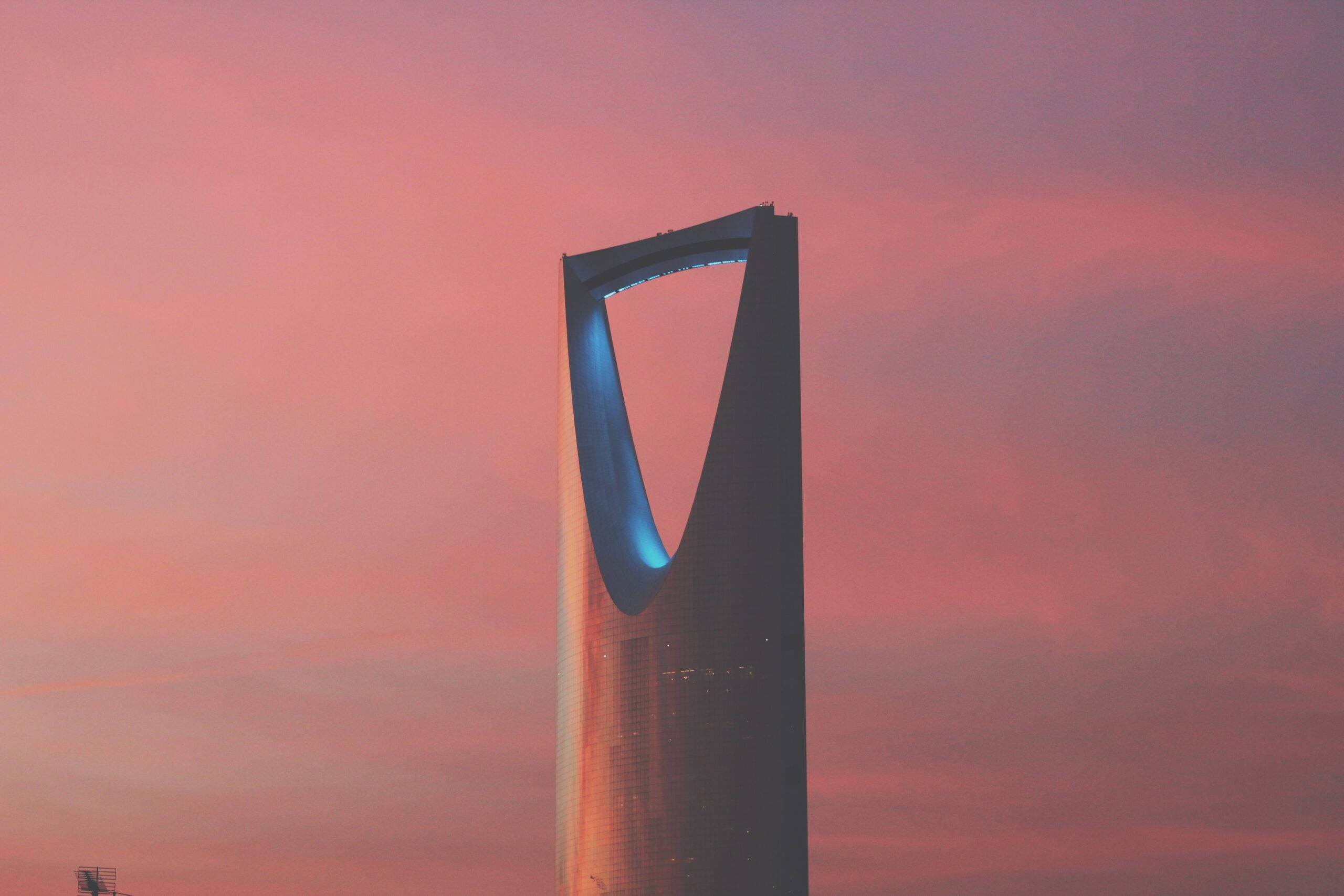 Saudi 桌布 壁紙 — 王國塔高聳入雲，映襯著充滿活力的粉紅色和橙色日落天空，其標誌性的藍色拱門閃閃發光。