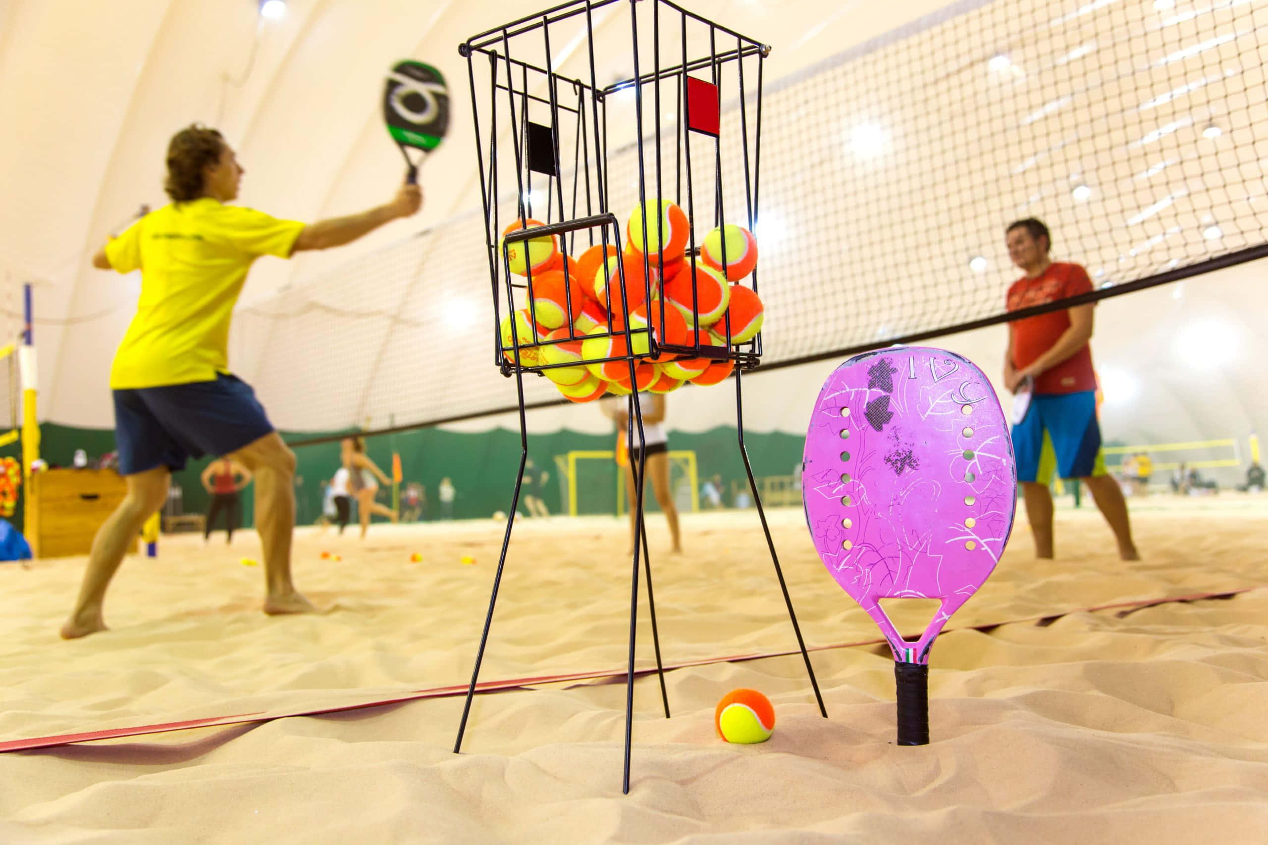 Beach Tennis 桌布 壁紙 — 玩家在室內沙灘匹克球比賽中使用紫色球拍和裝滿橙色球的球分配器。