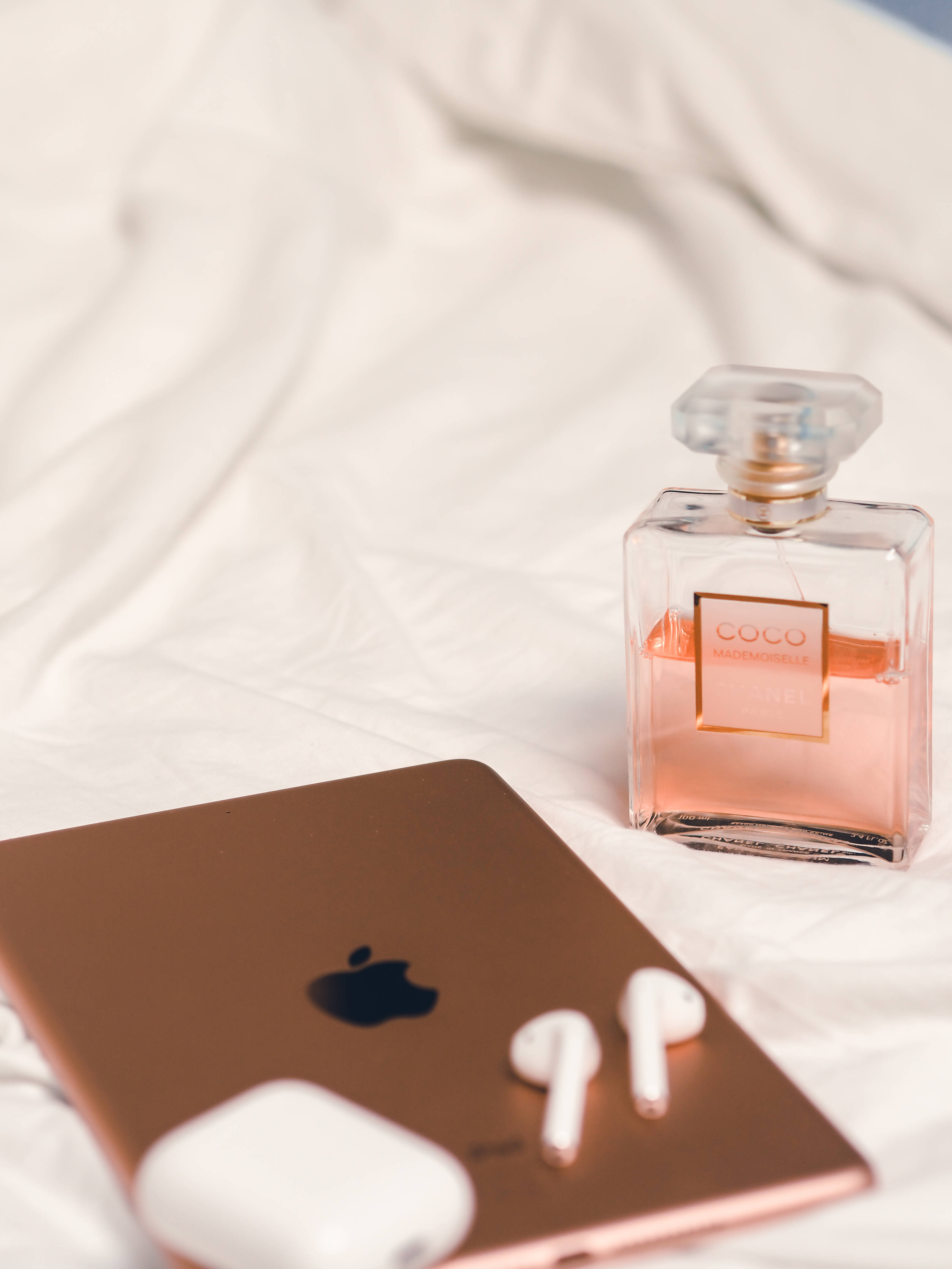 女孩感手機 桌布 壁紙 — 玫瑰金 iPhone 和白色 AirPods 放在一個粉紅色 Chanel Coco Mademoiselle 香水瓶旁邊，擱在皺巴巴的白色床單上。