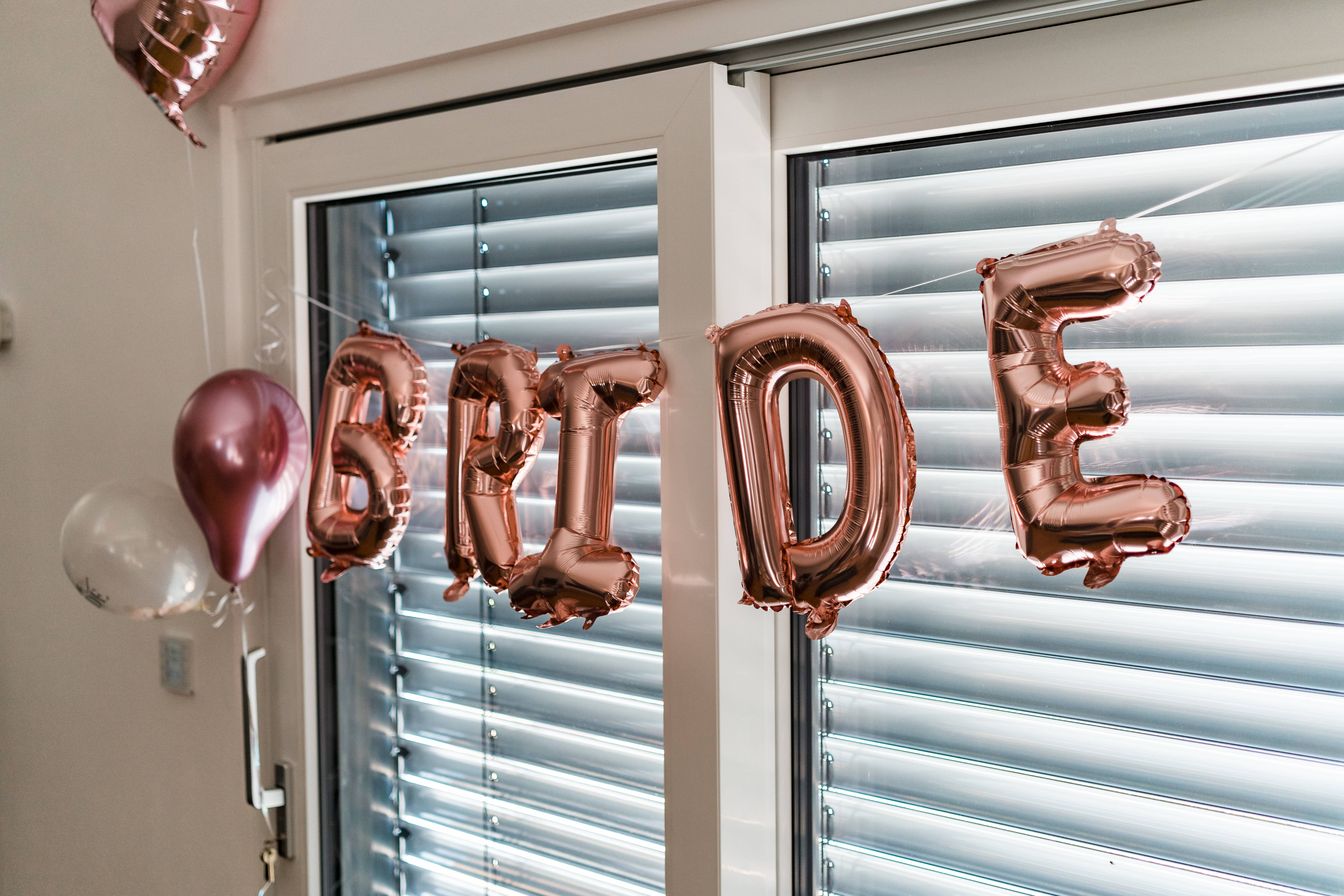 Bachelorette 桌布 壁紙 — 玫瑰金色箔氣球拼出 BRIDE 字樣，掛在窗簾關閉的窗戶附近，房間光線柔和。