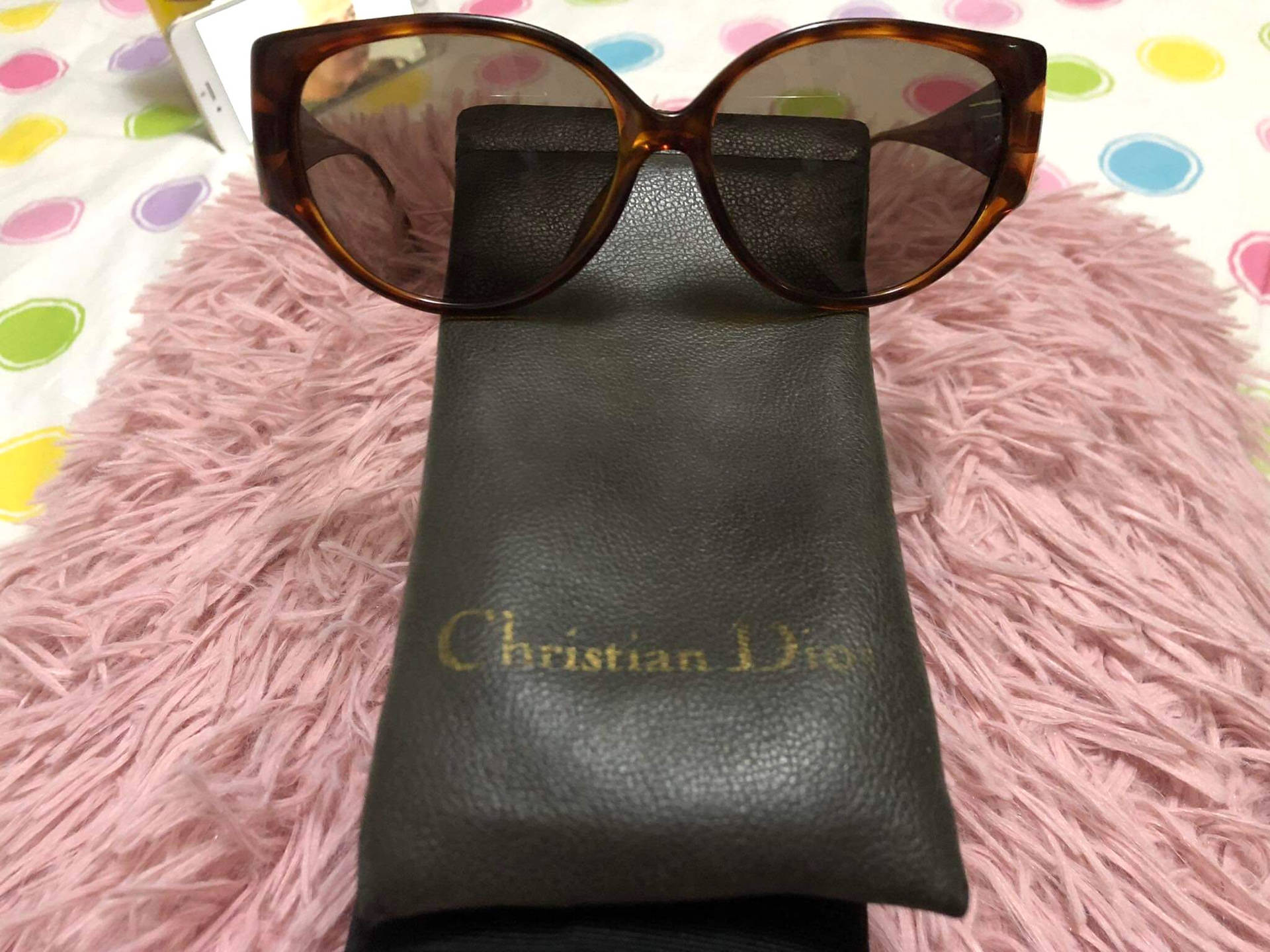 玳瑁色太陽眼鏡放在 Dior 眼鏡盒上，置於有圓點的粉紅色毛絨地毯上。 — Dior 動態 壁紙