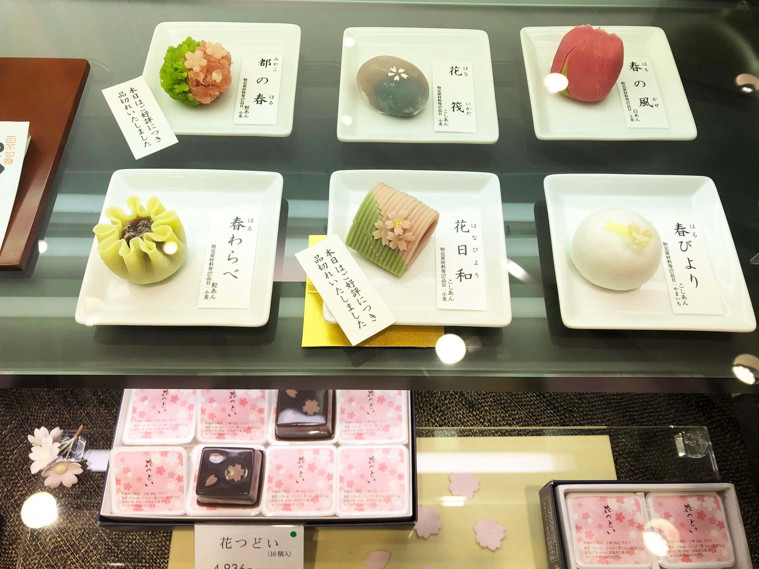 Japanese Sweets 桌布 壁紙 — 玻璃展示櫃中，白色盤子上擺放著精緻的日本和菓子，附有手寫標籤和櫻花圖案。