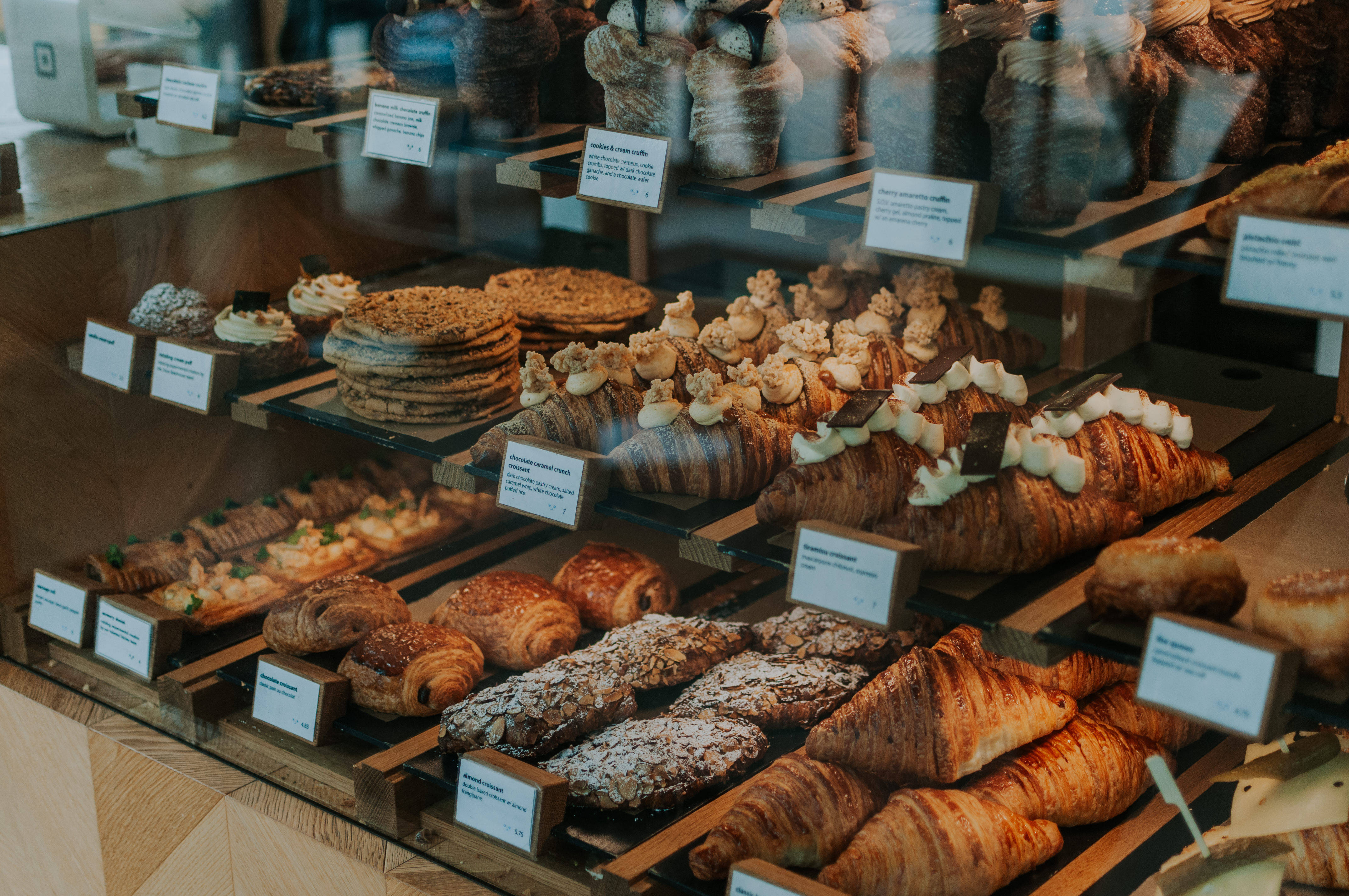 Pastries 桌布 壁紙 — 玻璃展示櫃內放滿金黃色的可頌、杏仁糕點和千層蛋糕，整齊排列在木架上，附有白色小標籤。