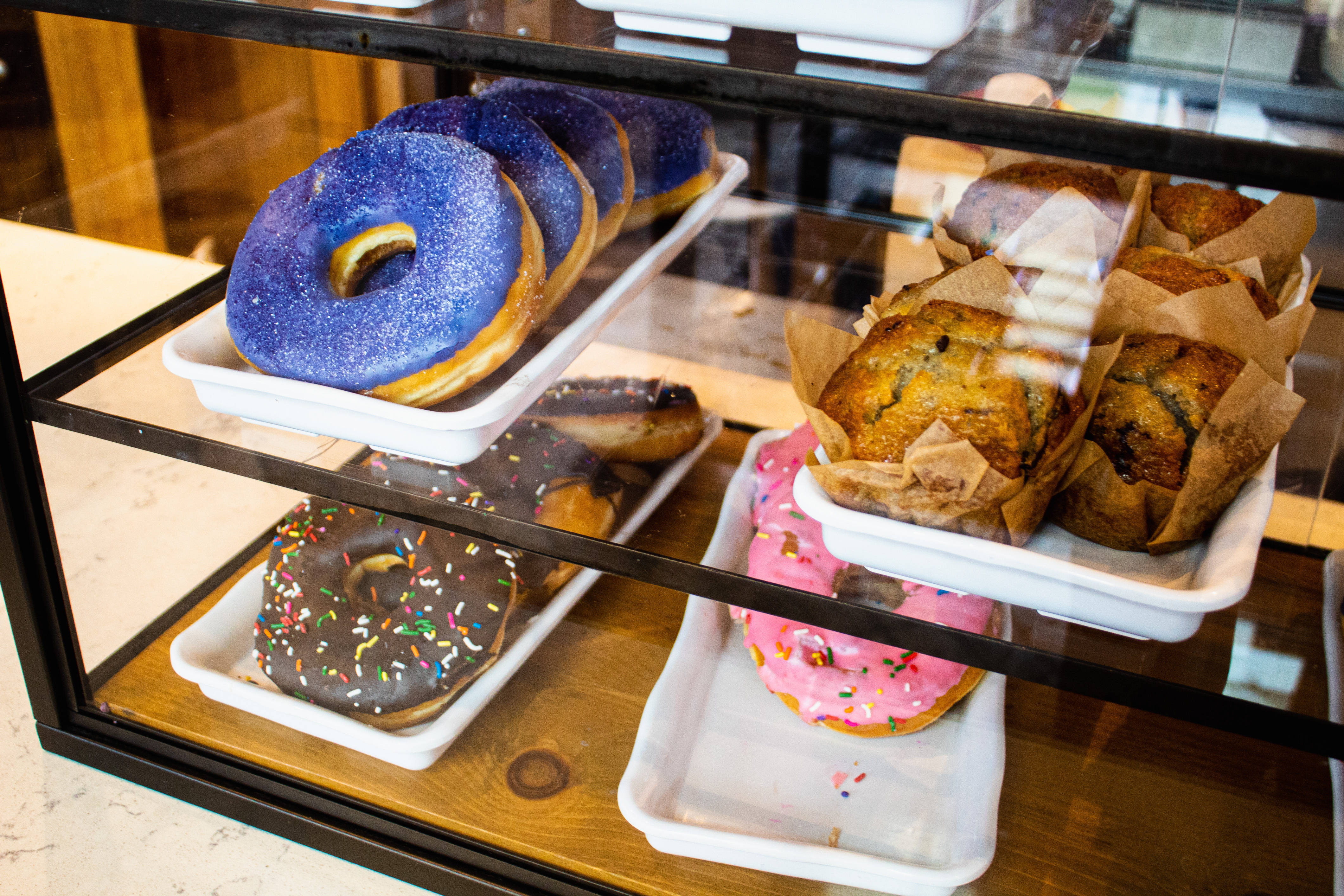 Pastries 桌布 壁紙 — 玻璃展示櫃內放著一盤盤色彩繽紛的甜甜圈和馬芬，上面有鮮豔的糖霜和彩針。