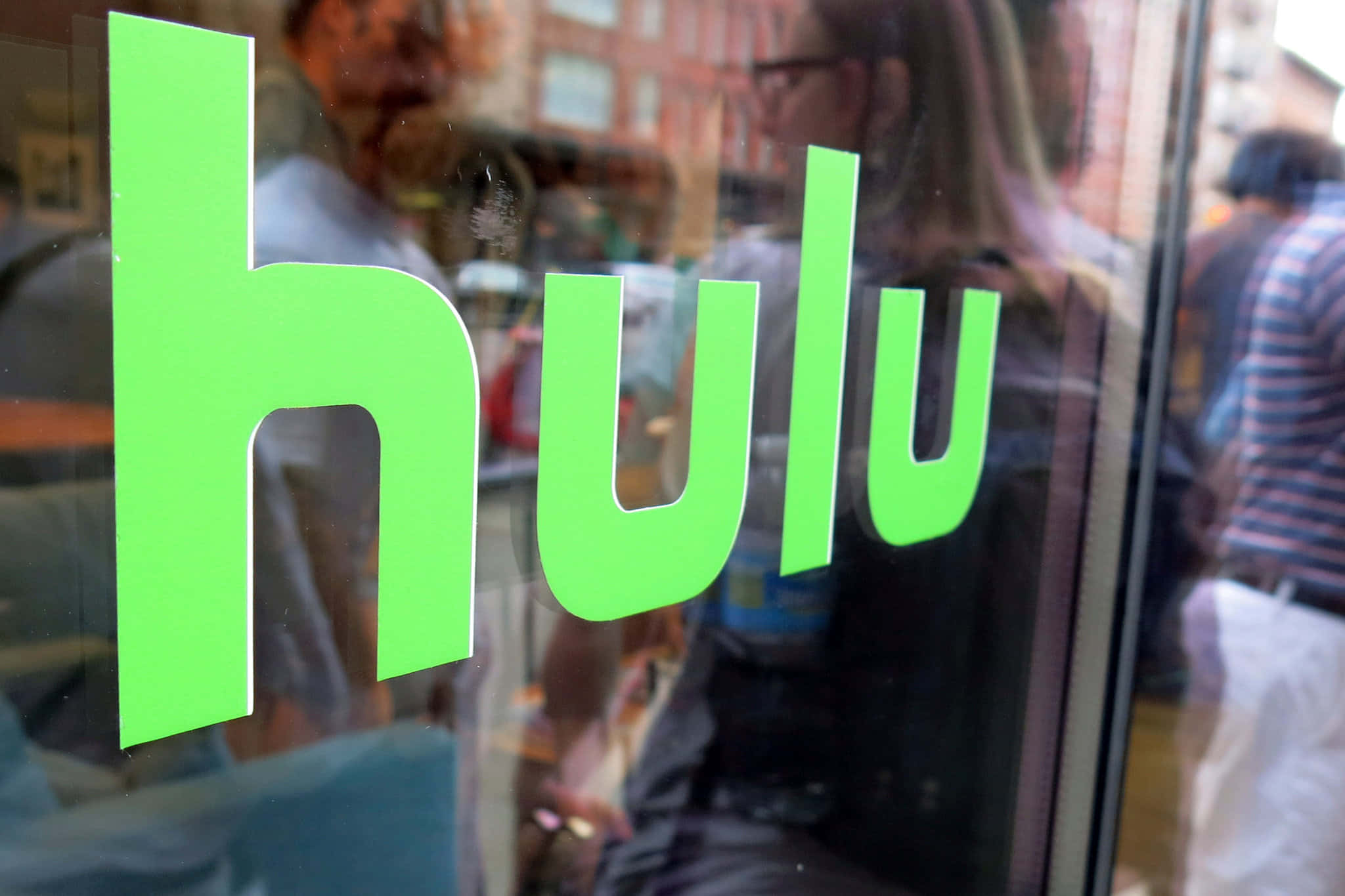 Hulu 桌布 壁紙 — 玻璃店面上鮮豔的綠色Hulu標誌，帶有模糊的行人和磚建築倒影。