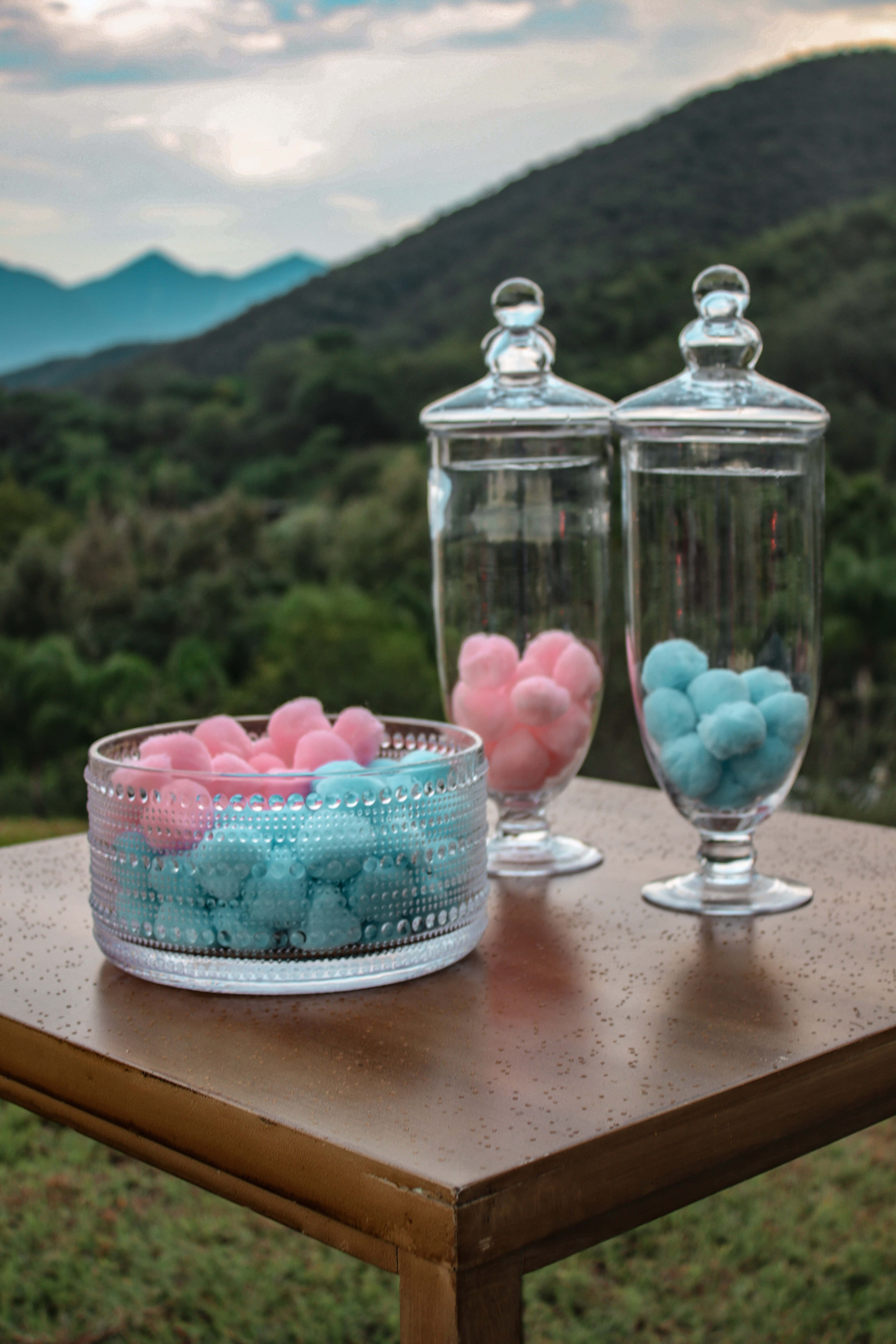 Baby Shower 桌布 壁紙 — 玻璃罐和碗裝滿粉紅色和藍色的棉花球，放在木桌上，背景是鬱鬱蔥蔥的綠色山脈，在柔和的暮色天空下。