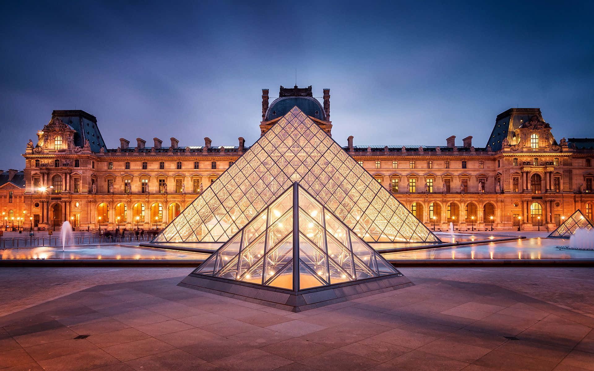 Paris Desktop 桌布 壁紙 — 玻璃金字塔在黃昏時分溫暖地發光，映襯著歷史悠久的羅浮宮博物館外牆，噴泉反射著金色的光芒。