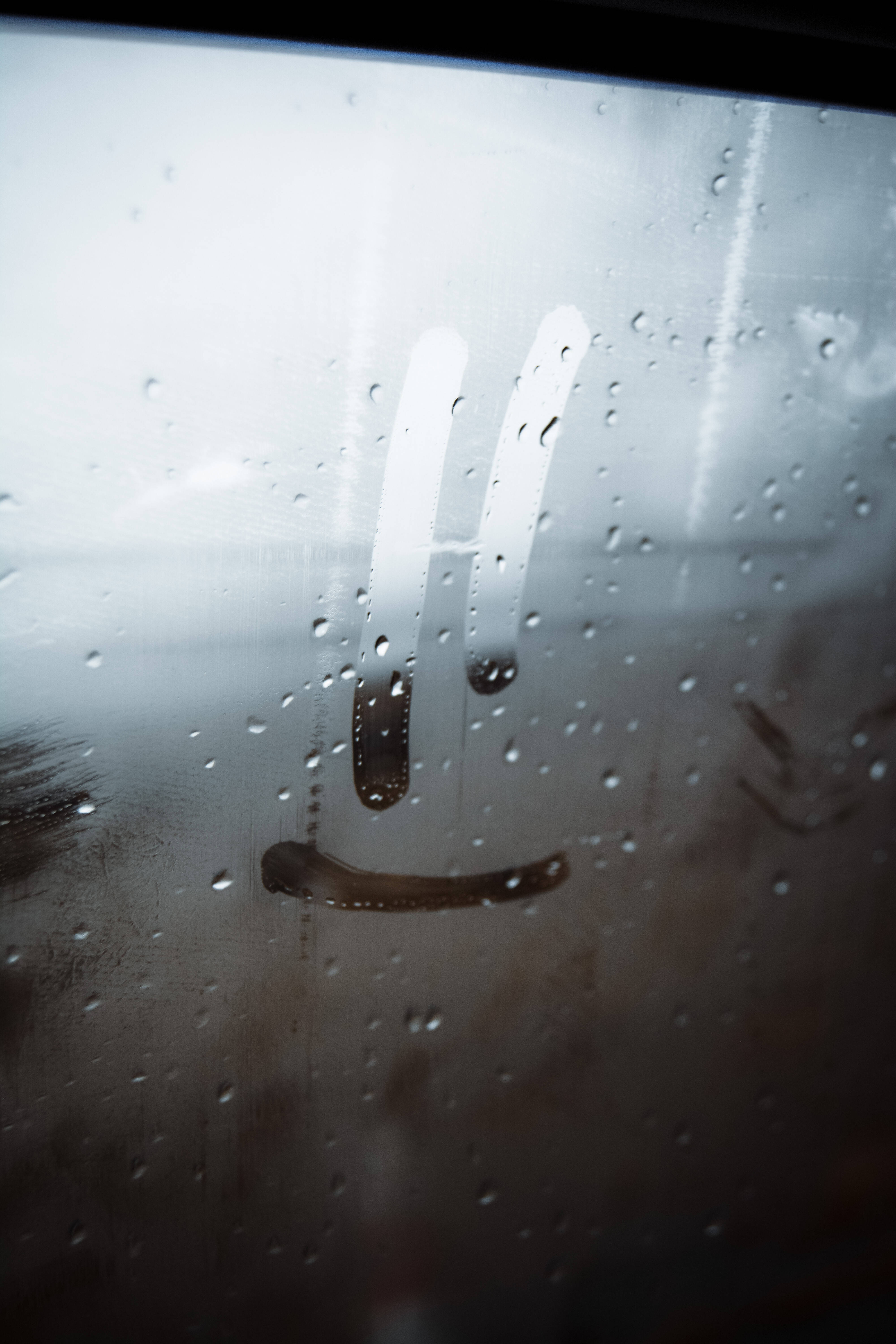 鎖屏 桌布 壁紙 — 玻璃門上雨滴的特寫，上面畫著一個模糊的笑臉，營造出一種陰鬱而內省的氛圍。