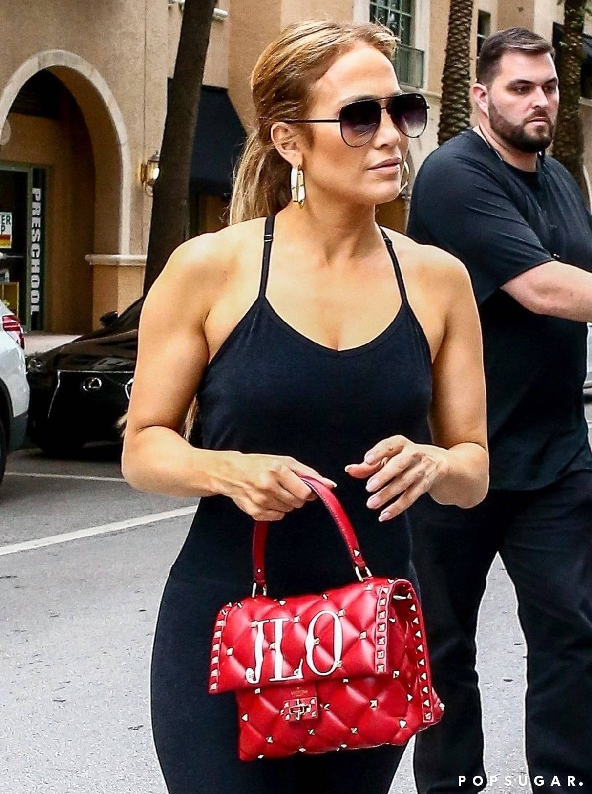 Celebrity 桌布 壁紙 — 珍妮佛洛佩茲自信地穿著黑色連衣裙，在陽光明媚的城市街道上拿著一個大膽的紅色鉚釘JLO手提包。