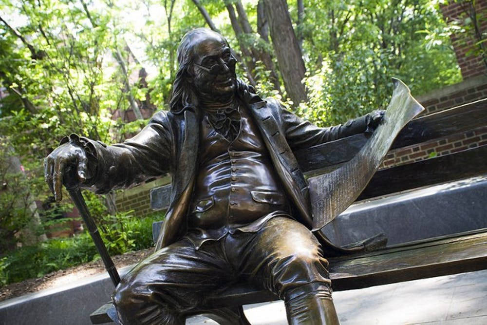 Franklin 桌布 壁紙 — 班傑明·富蘭克林的銅像放鬆地坐在長椅上，手持報紙和手杖，周圍環繞著茂密的綠樹。