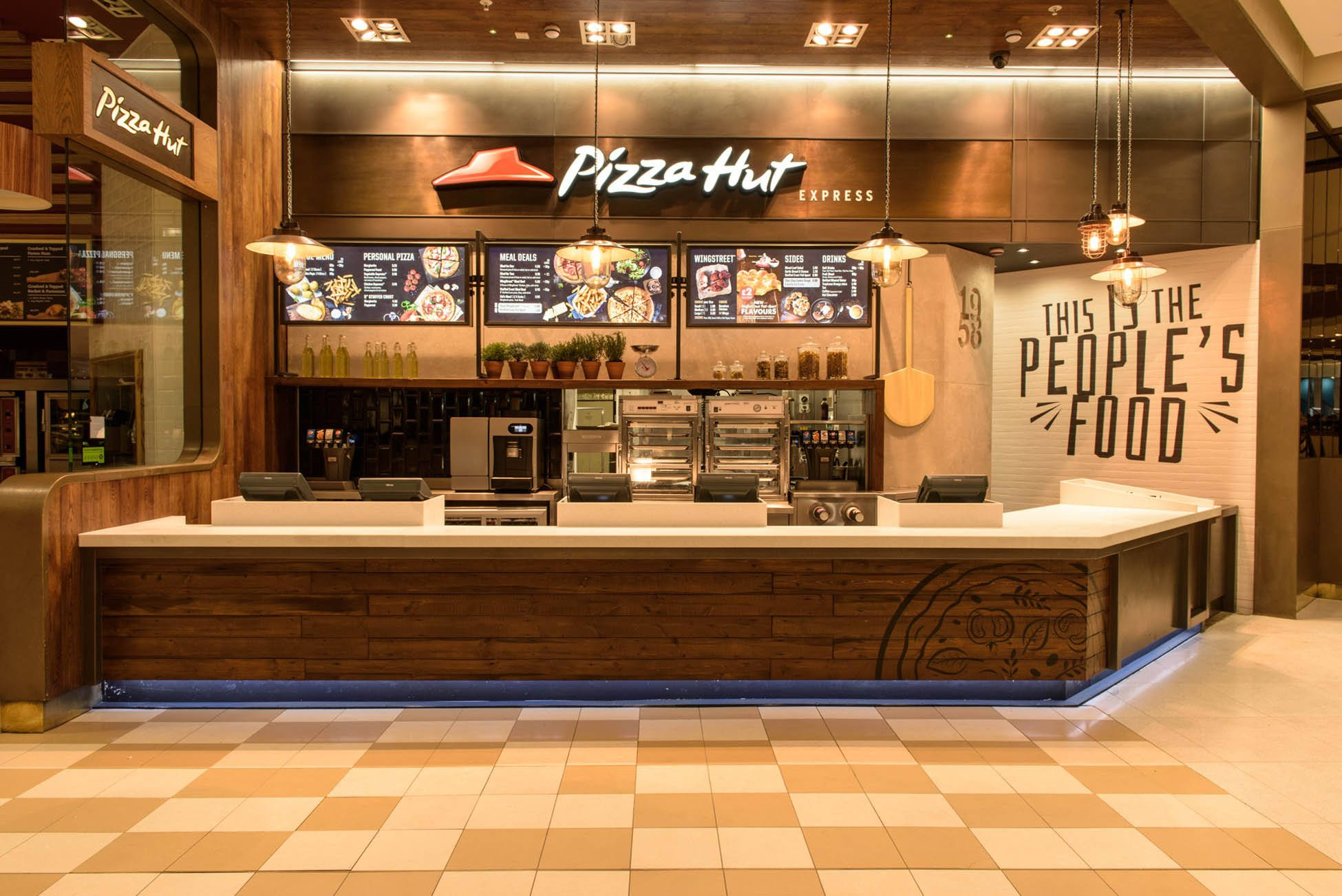 Hut 桌布 壁紙 — 現代 Pizza Hut Express 餐廳內部，設有木製櫃檯、數位菜單，方格地板上有溫暖的吊燈。
