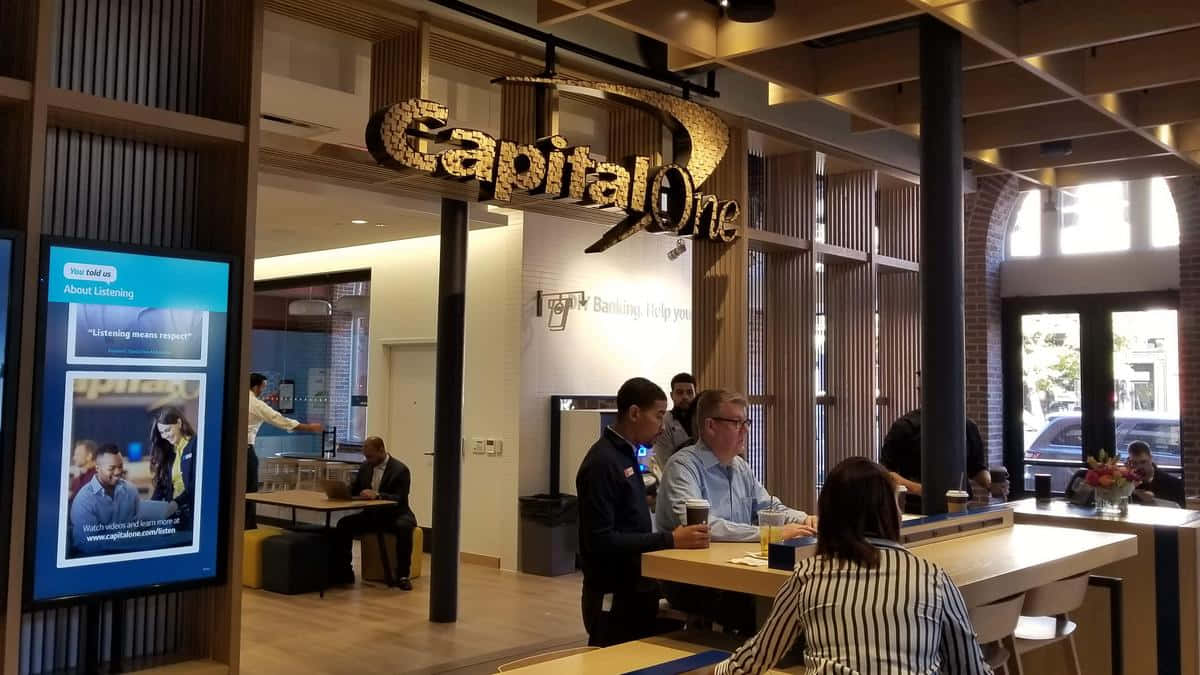 咖啡動圖 桌布 壁紙 — 現代Capital One銀行內部，顧客和員工在溫暖燈光下於時尚木桌互動。
