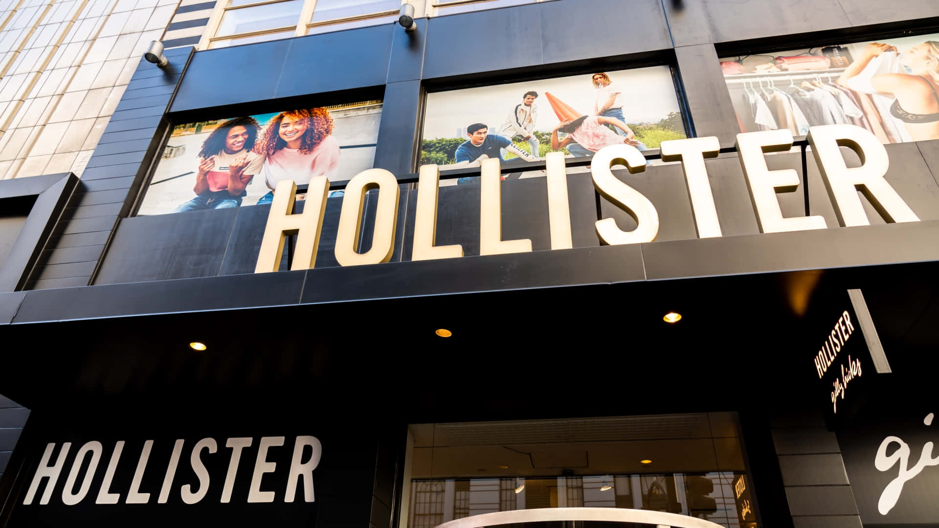 Hollister 桌布 壁紙 — 現代Hollister商店外牆，帶有醒目的白色招牌和大型窗戶中充滿活力的生活方式廣告。