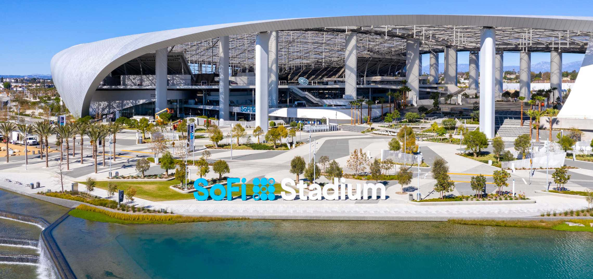 Sofi Stadium 桌布 壁紙 — 現代SoFi Stadium的鳥瞰圖，在晴朗的藍天下，前景有棕櫚樹和藍綠色水景。