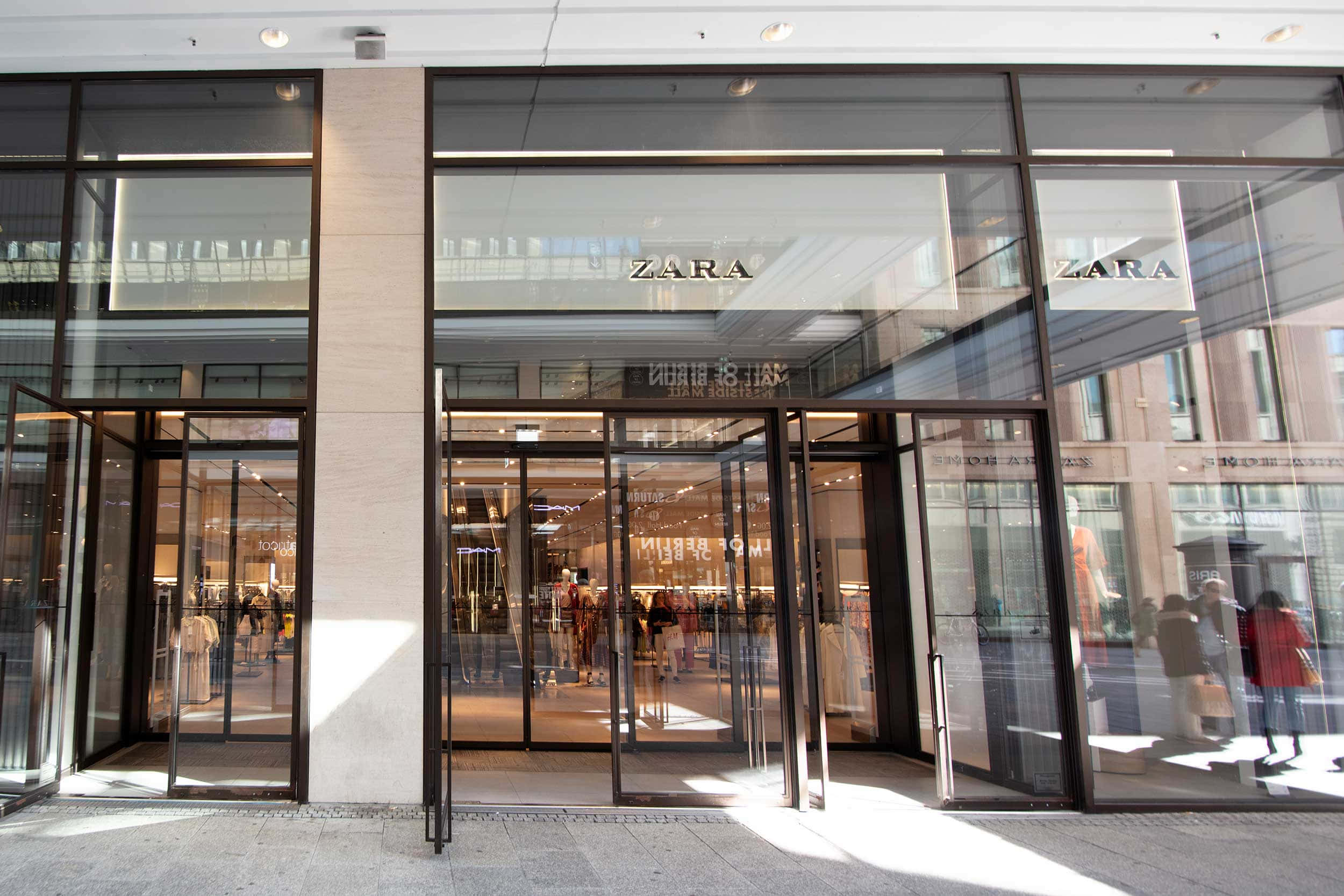 Zara 桌布 壁紙 — 現代Zara店鋪外牆，設有玻璃門和反光窗戶，在明亮日光下顯示店內的購物者。
